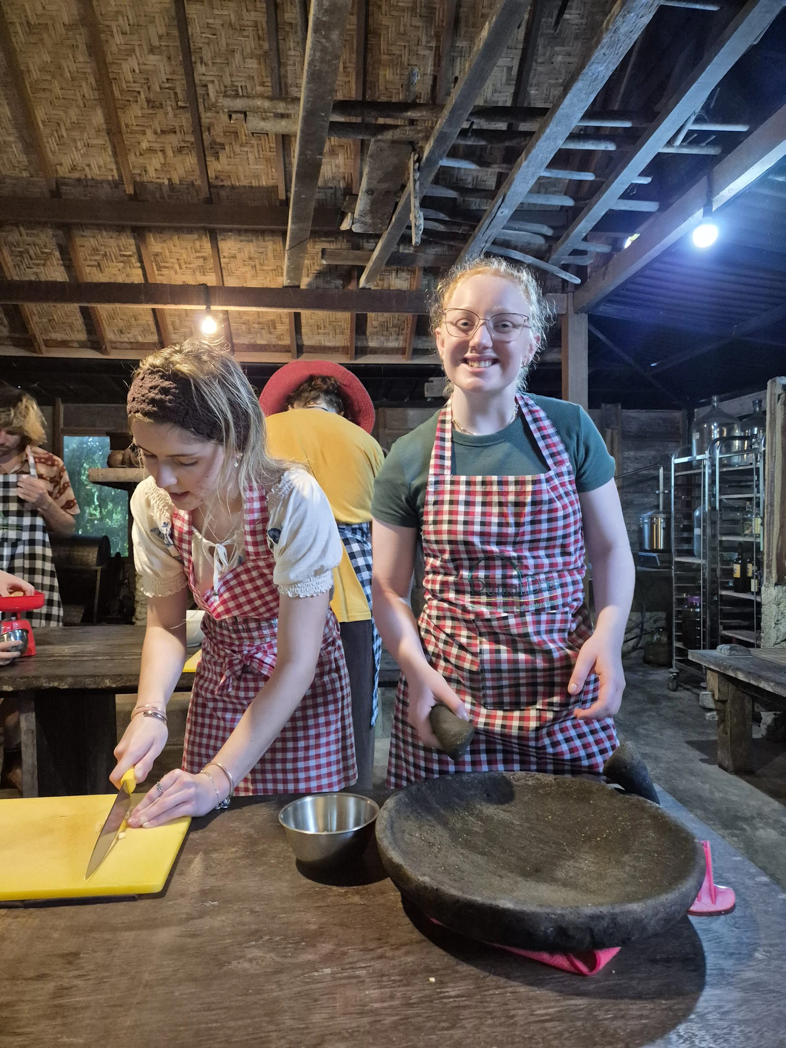 Two teen travelers in checked aprons smile as they chop and grind ingredients with a stone mortar at a wooden table in a Bali kitchen