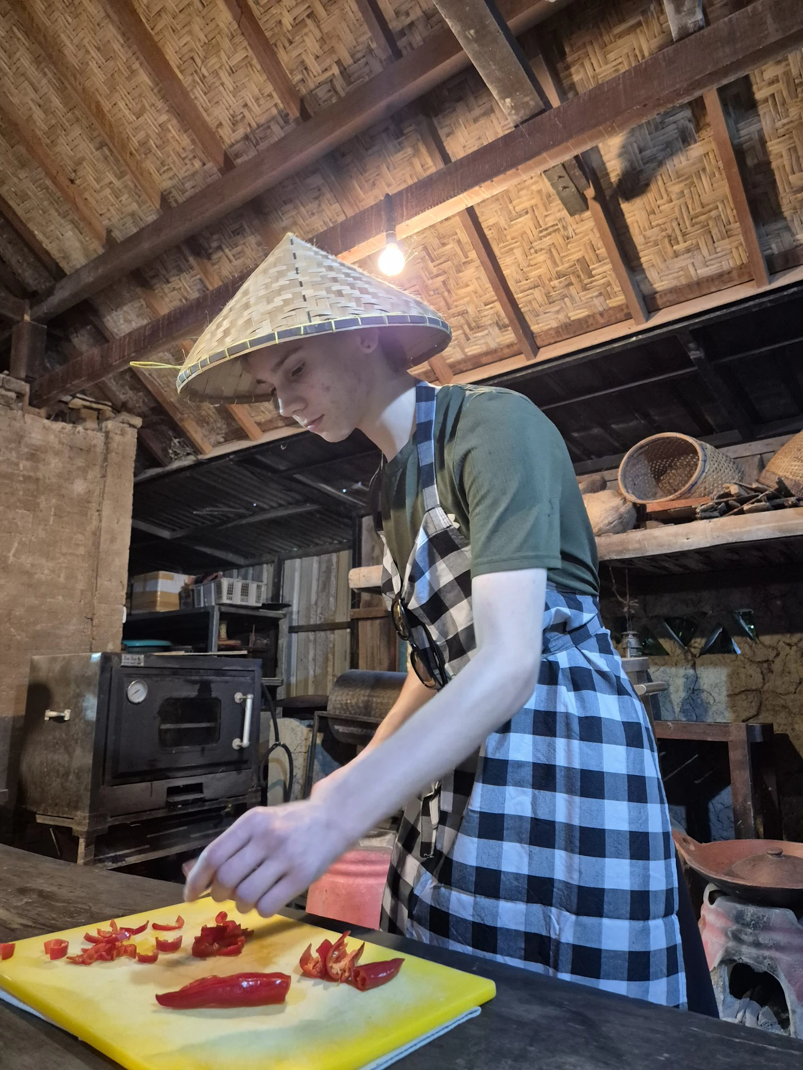 A teen traveler in a conical straw hat and checked apron chops red chilies on a yellow board in a rustic wooden kitchen in Bali