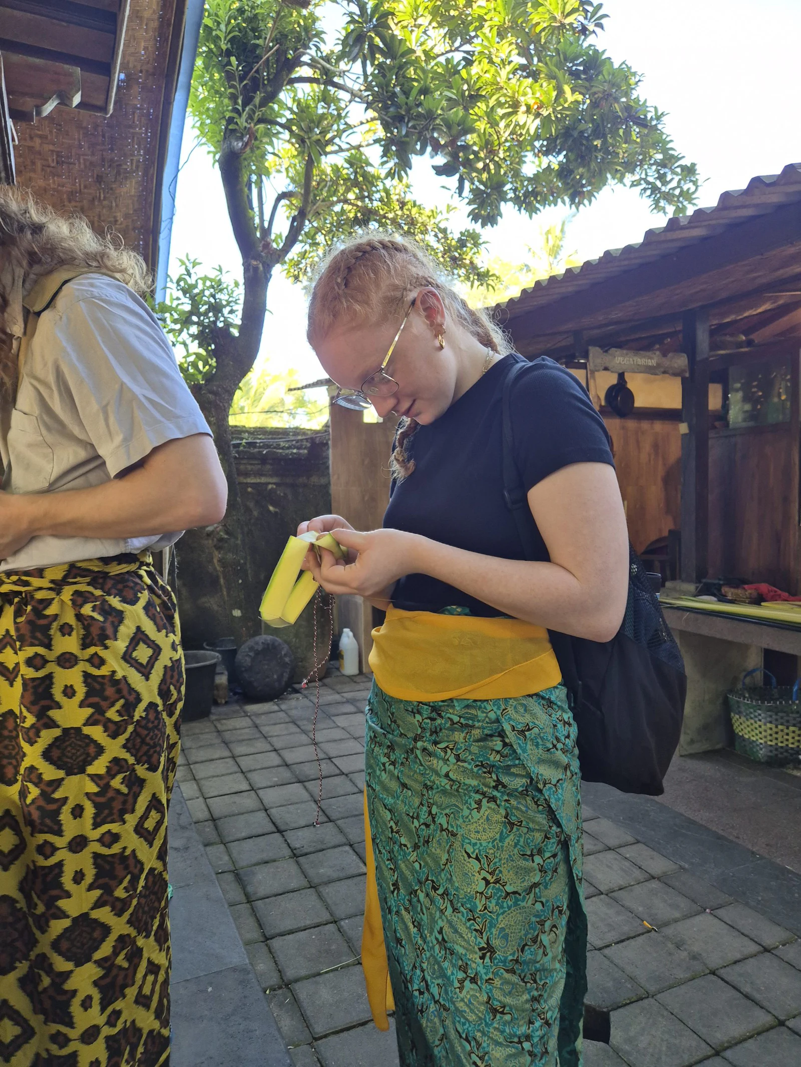 Two teen travelers in batik sarongs stand on a tiled courtyard in Bali, one looking down at a yellow leaf in her hands