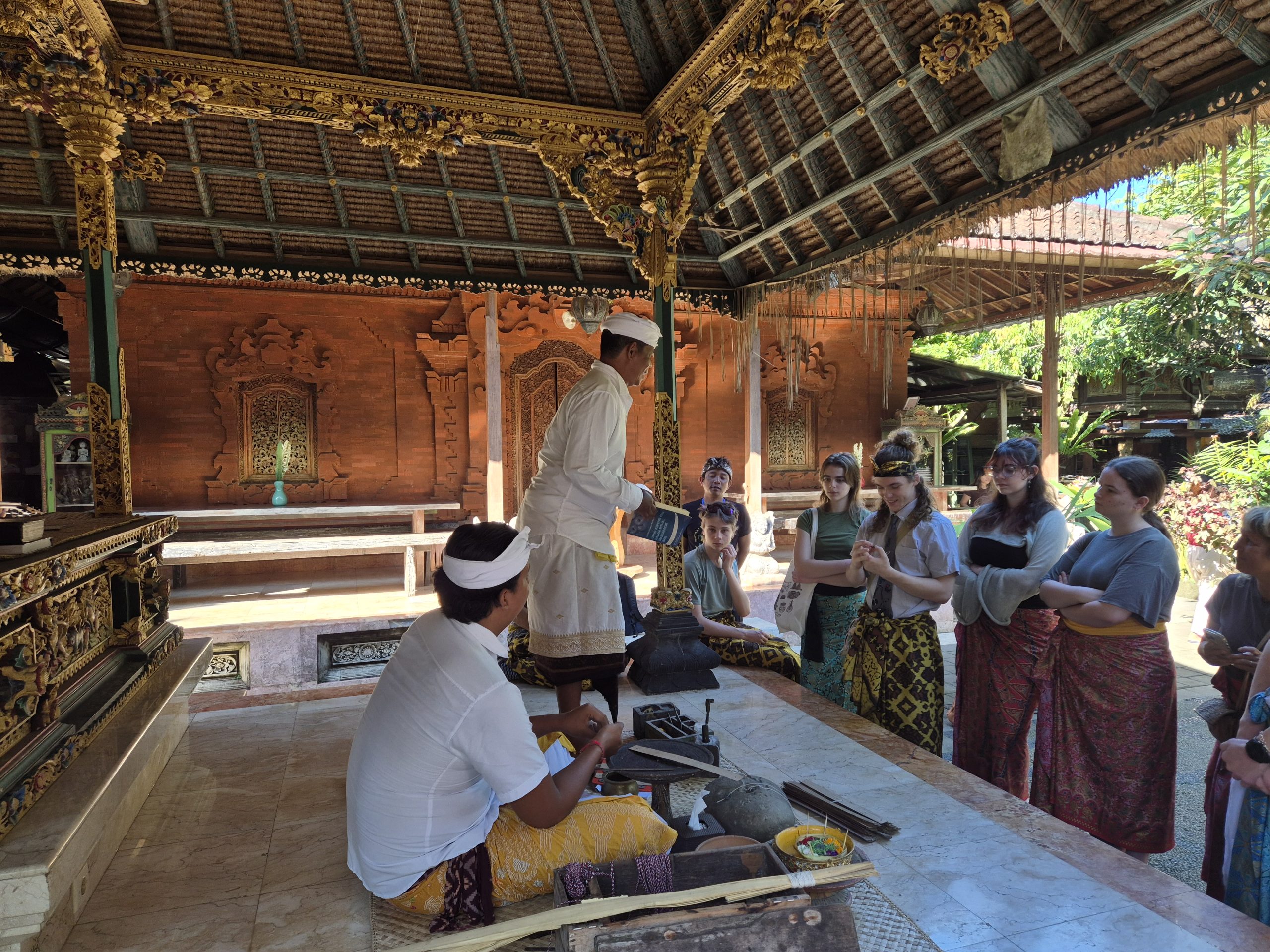 Students in sarongs gather in an ornate temple pavilion as Balinese hosts in white head wraps lead a demonstration