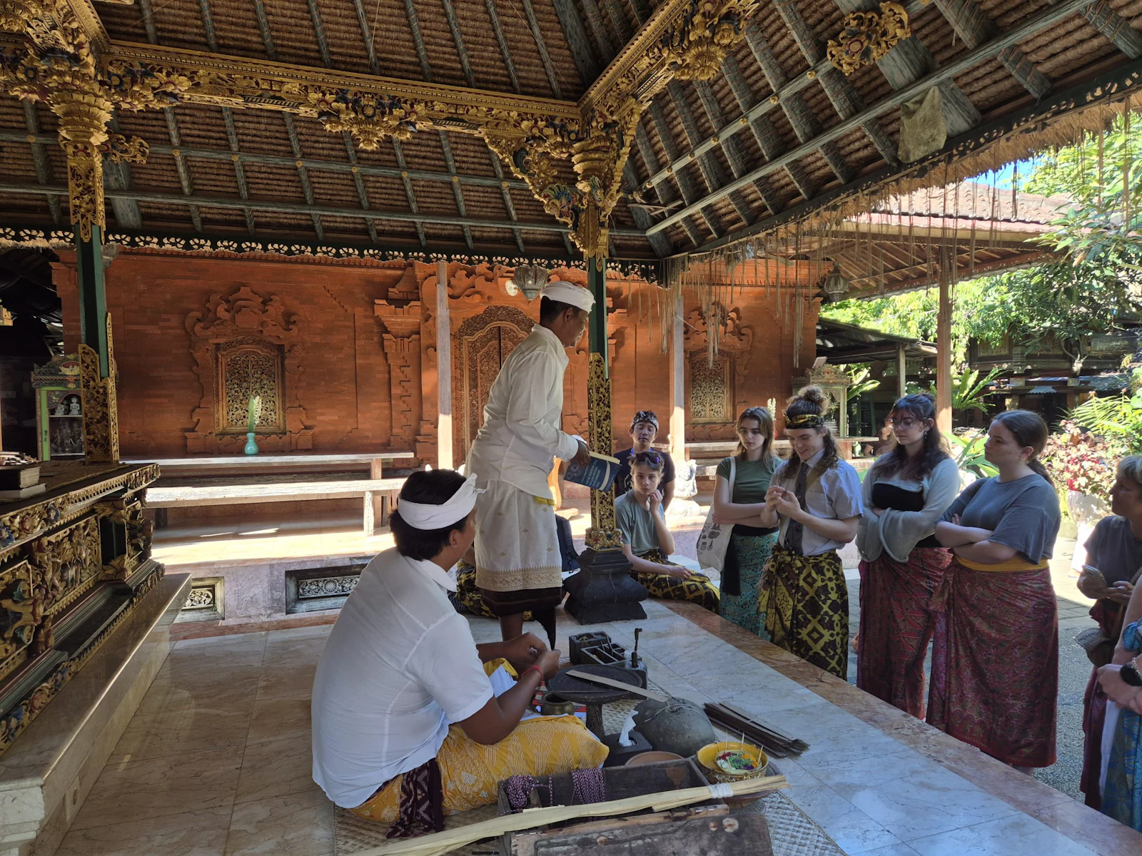 Teen travelers in batik sarongs stand at the back of an ornate carved wooden pavilion in Bali while a priest in white stands beside a seated man at a low table