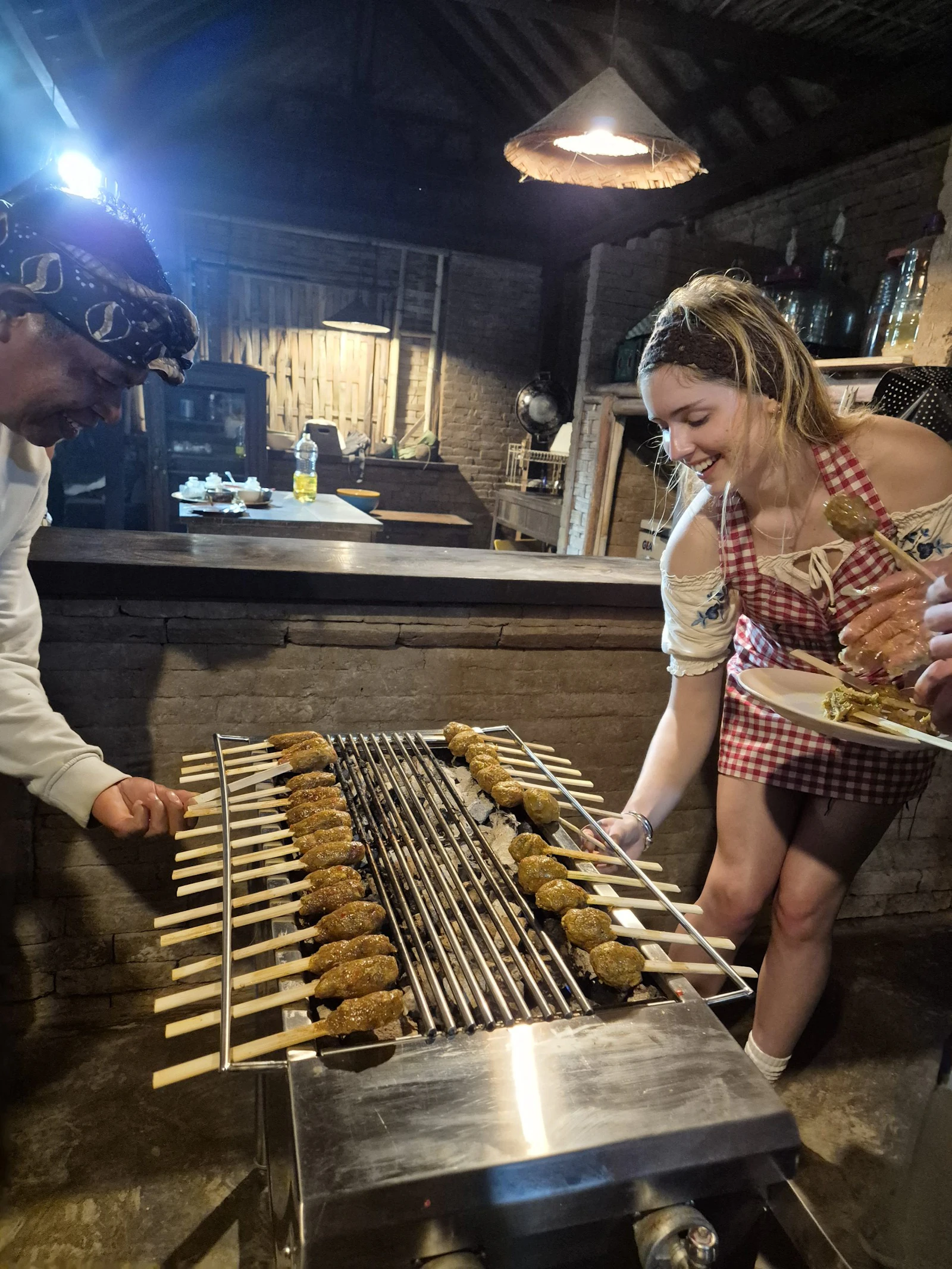 A teen traveler in a checked apron smiles as she turns rows of skewers on a charcoal grill in a dim wooden kitchen in Bali, a cook beside her