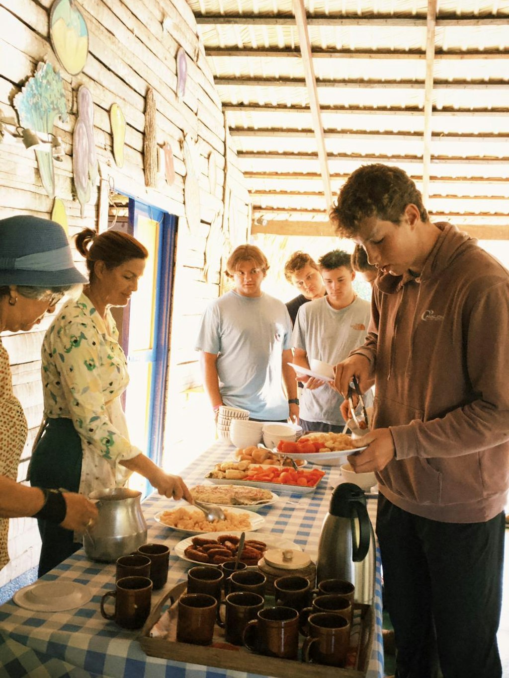 A group of young people lines up to enjoy a homemade meal served by local hosts in a rustic setting with wooden walls and a checkered tablecloth.