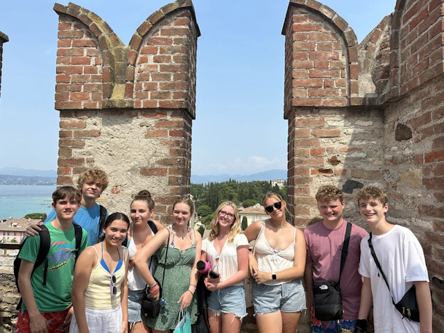 Teen travelers stand between the brick battlements of a castle tower in Italy, with a lake and hills behind them