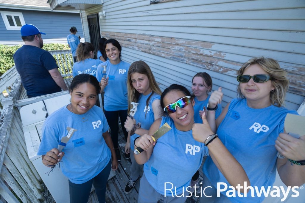 High school students in blue Rustic Pathways shirts holding paintbrushes and giving thumbs up while preparing for a service project.