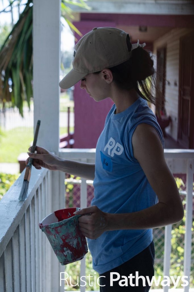 A student in a beige cap and Rustic Pathways shirt carefully painting porch railings during a service project.
