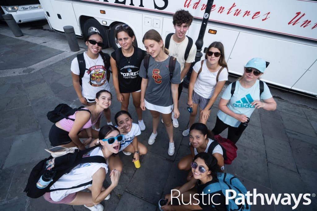 A group of high school students smiling and posing in front of a tour bus during a service learning trip to New Orleans.