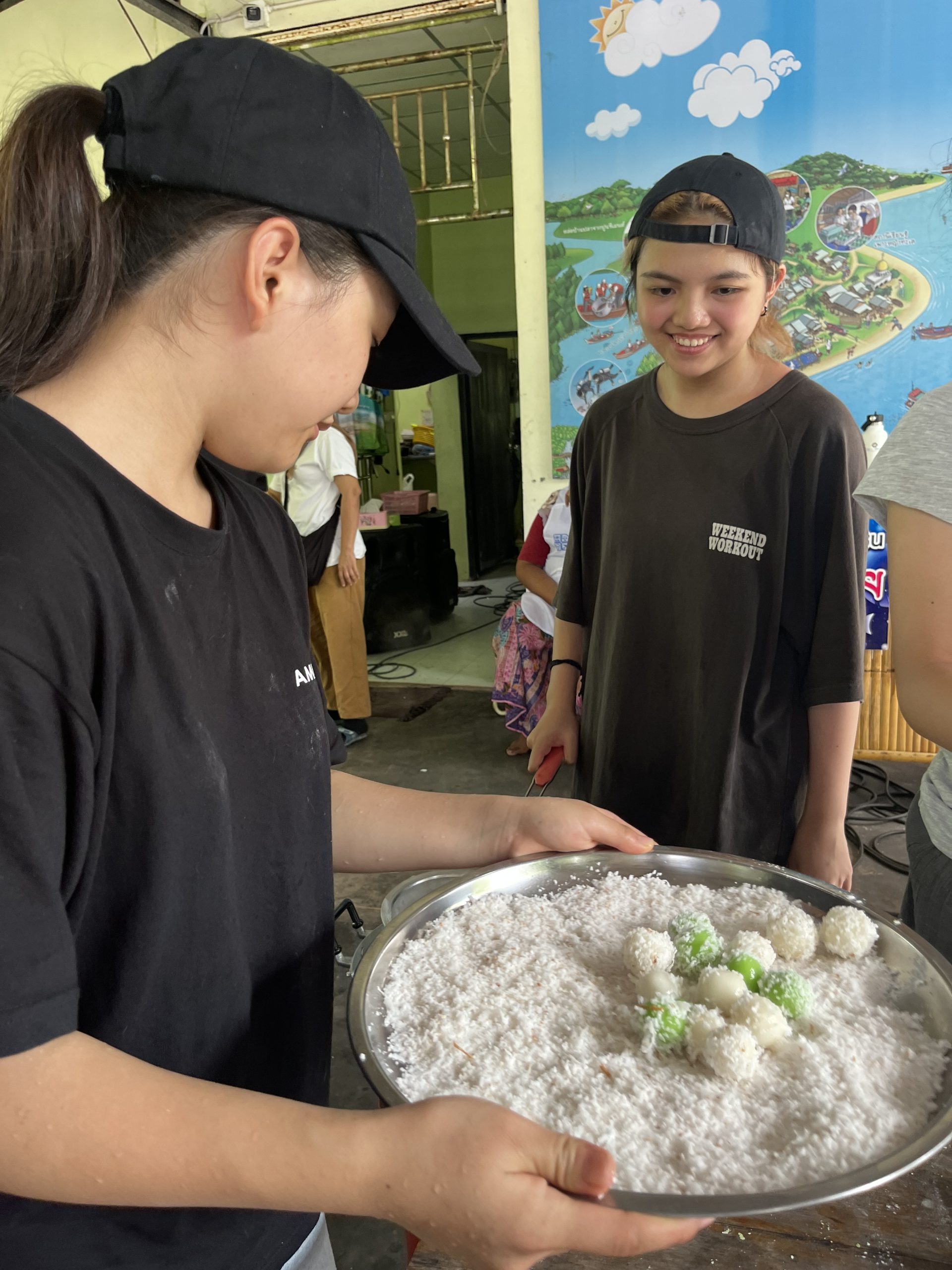 A student holds a tray of coconut-coated rice balls while another smiles nearby