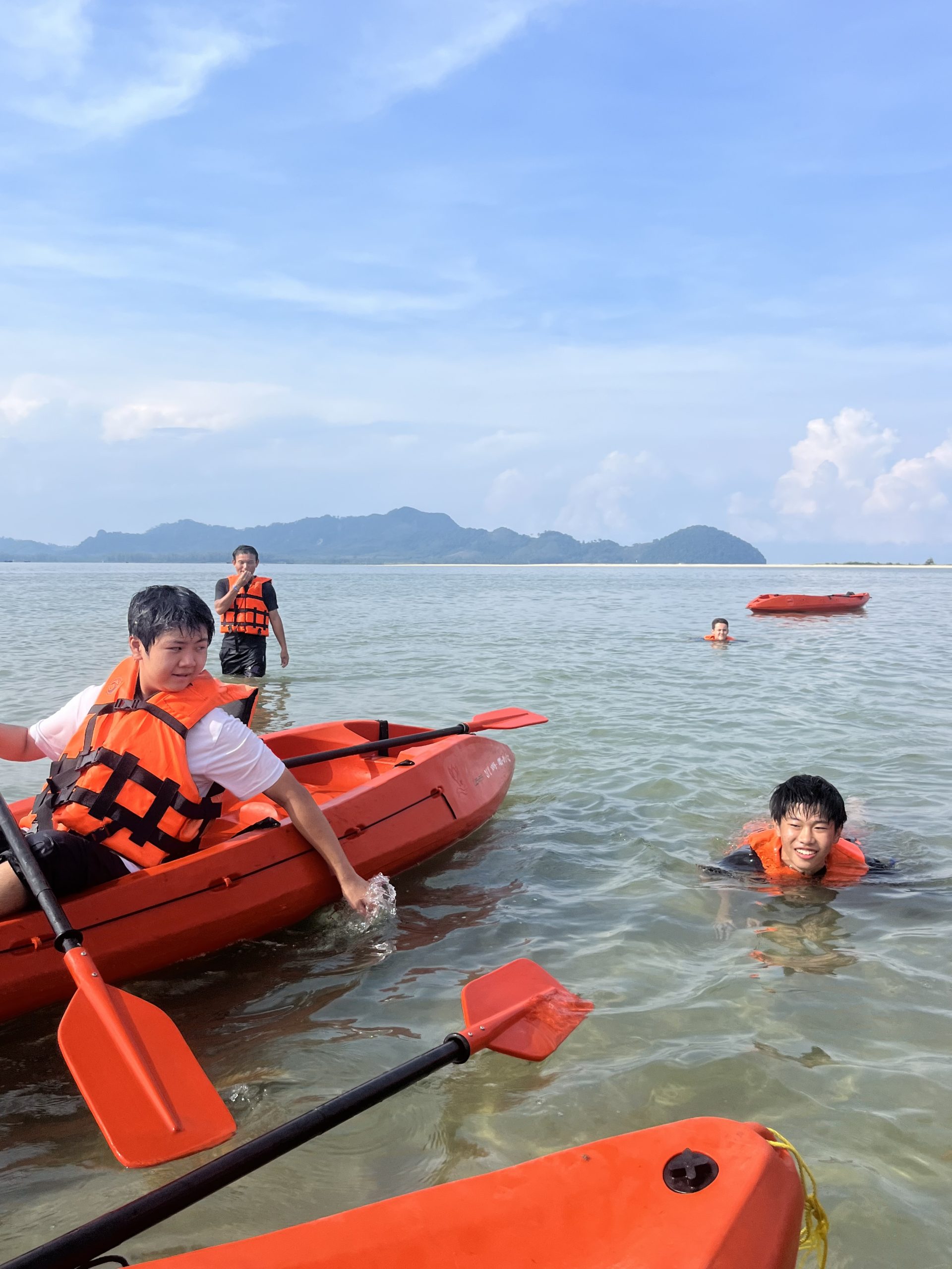 Students in life vests kayak and swim in shallow coastal waters with hills in the distance