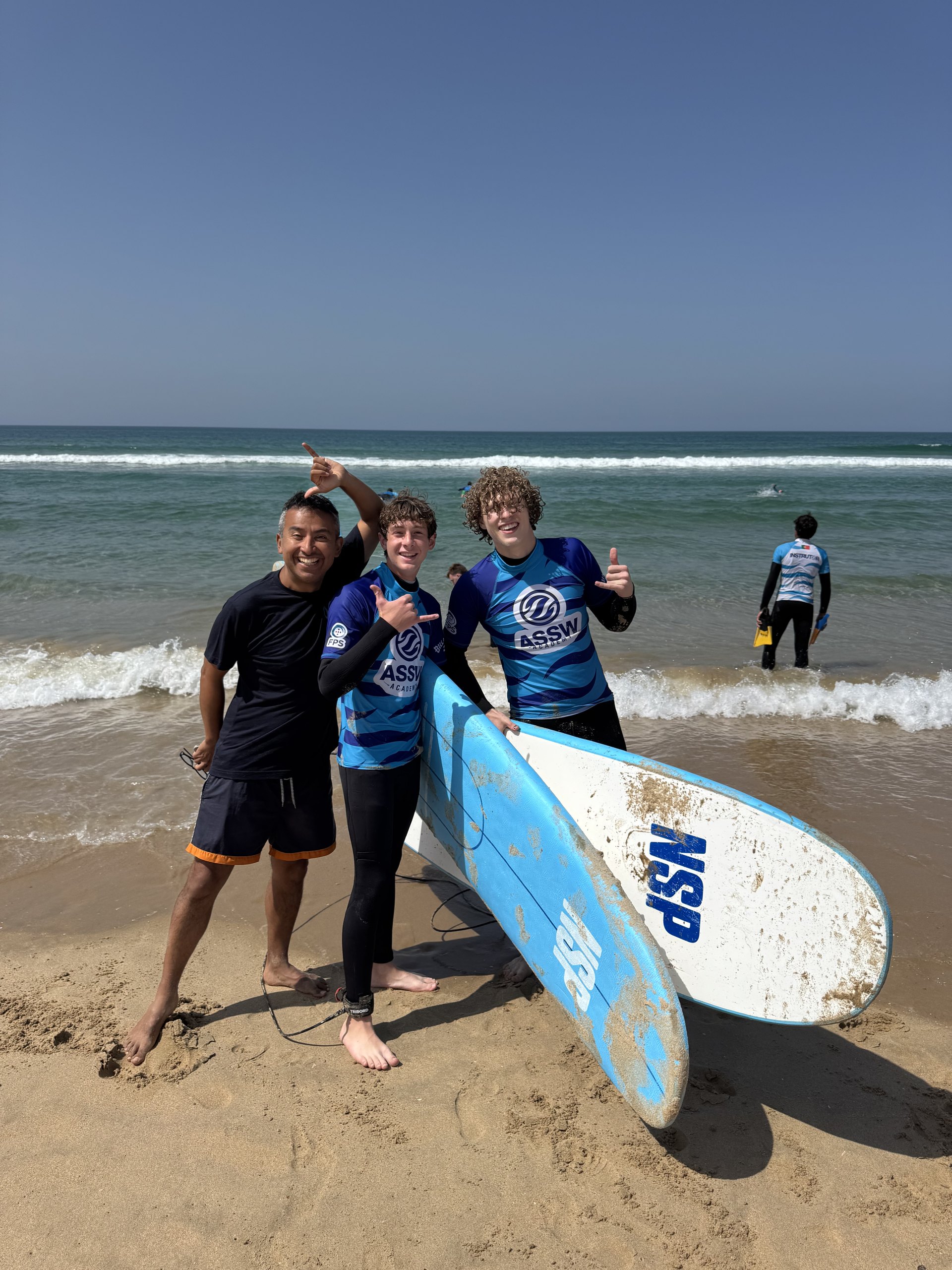 A surf instructor and two teen students in rash guards pose with surfboards at the water's edge