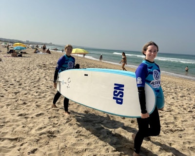 Two students carry a surfboard on a beach during a surf lesson on a teen tour in Portugal and Spain.