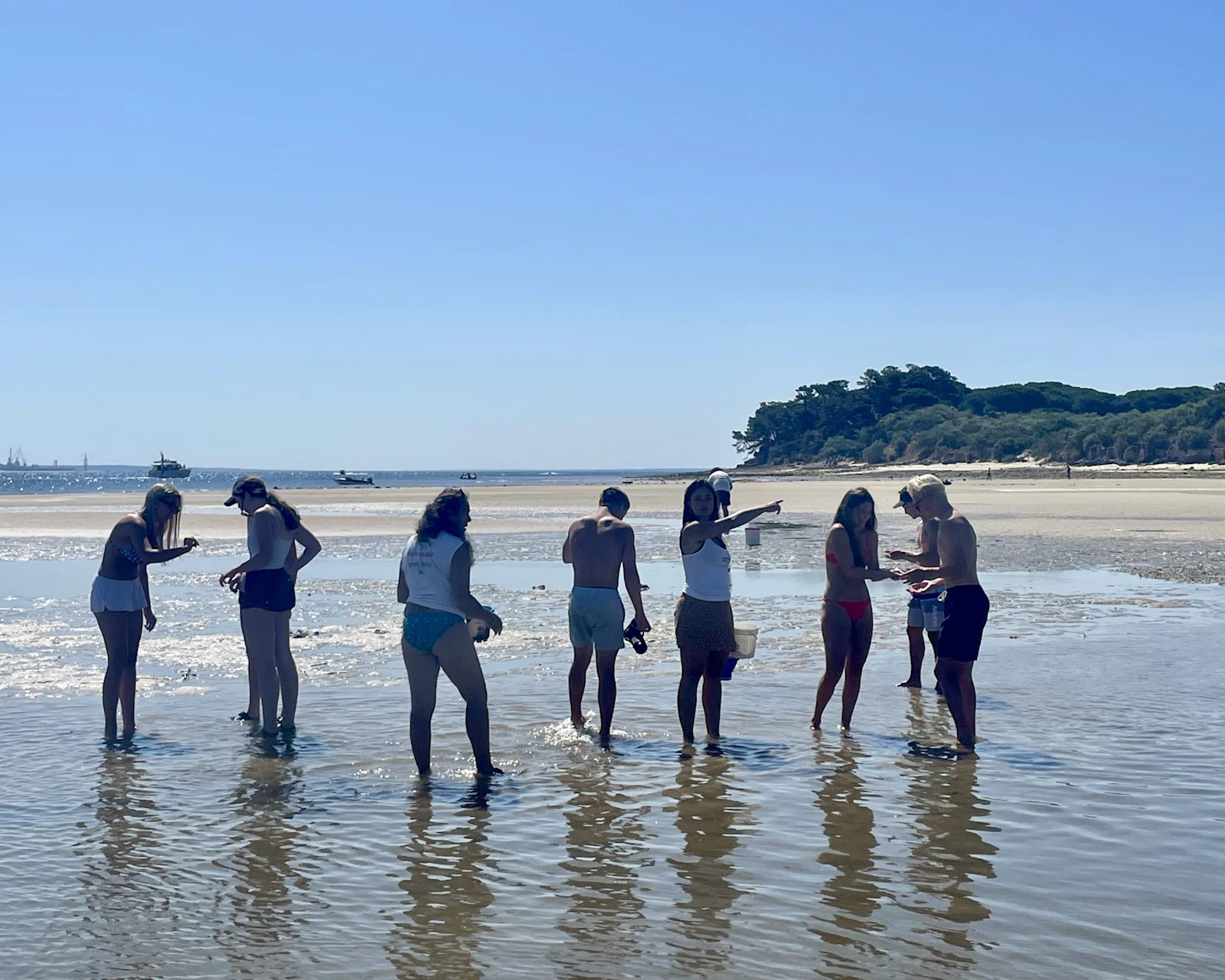 Teen travelers wade across a shallow tidal flat toward a wooded headland on the Portuguese coast