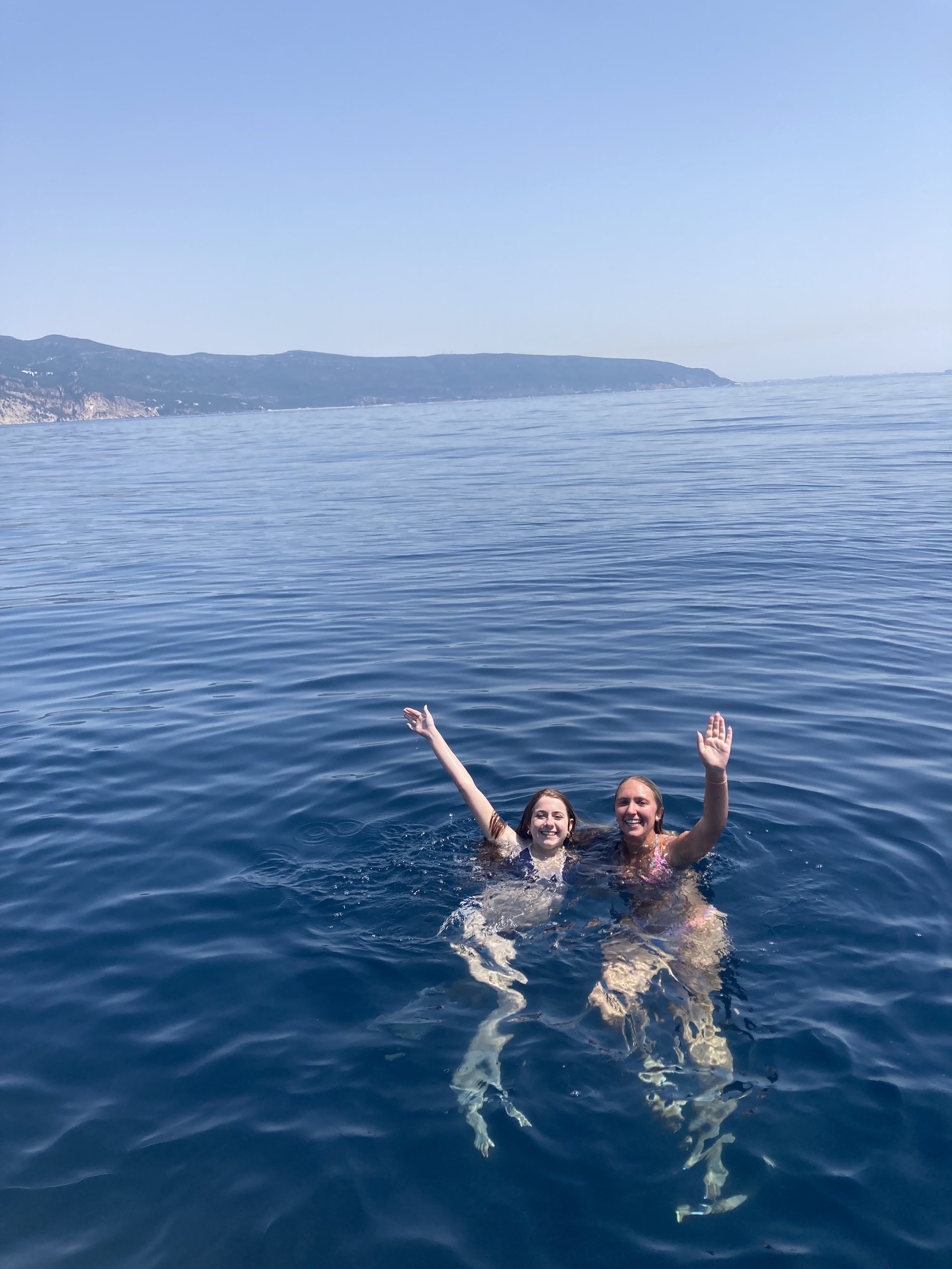 Two teen travelers wave while swimming in calm, deep blue sea off the coast