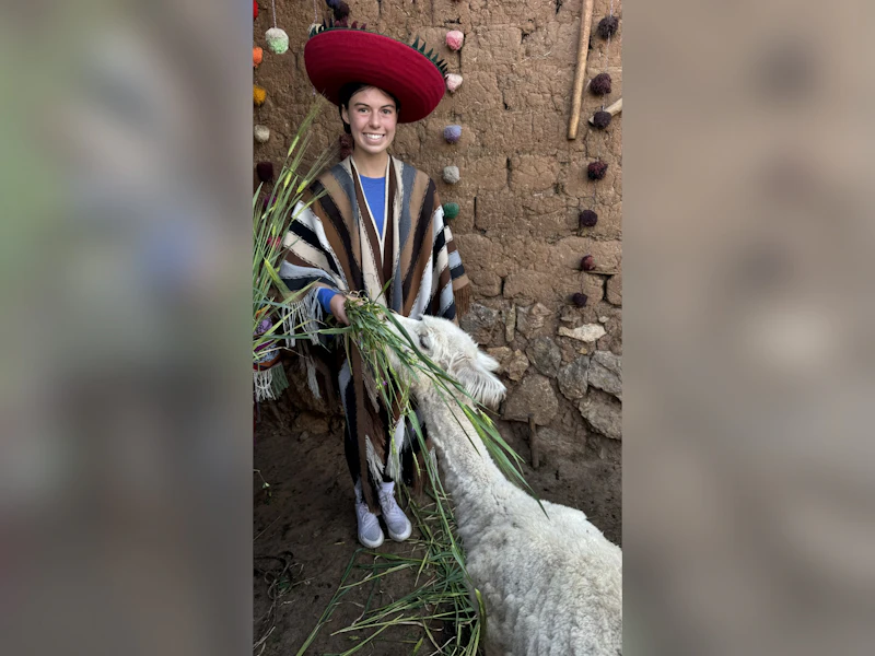A teen traveler in a red Andean hat and striped poncho feeds grass to a white alpaca in Peru