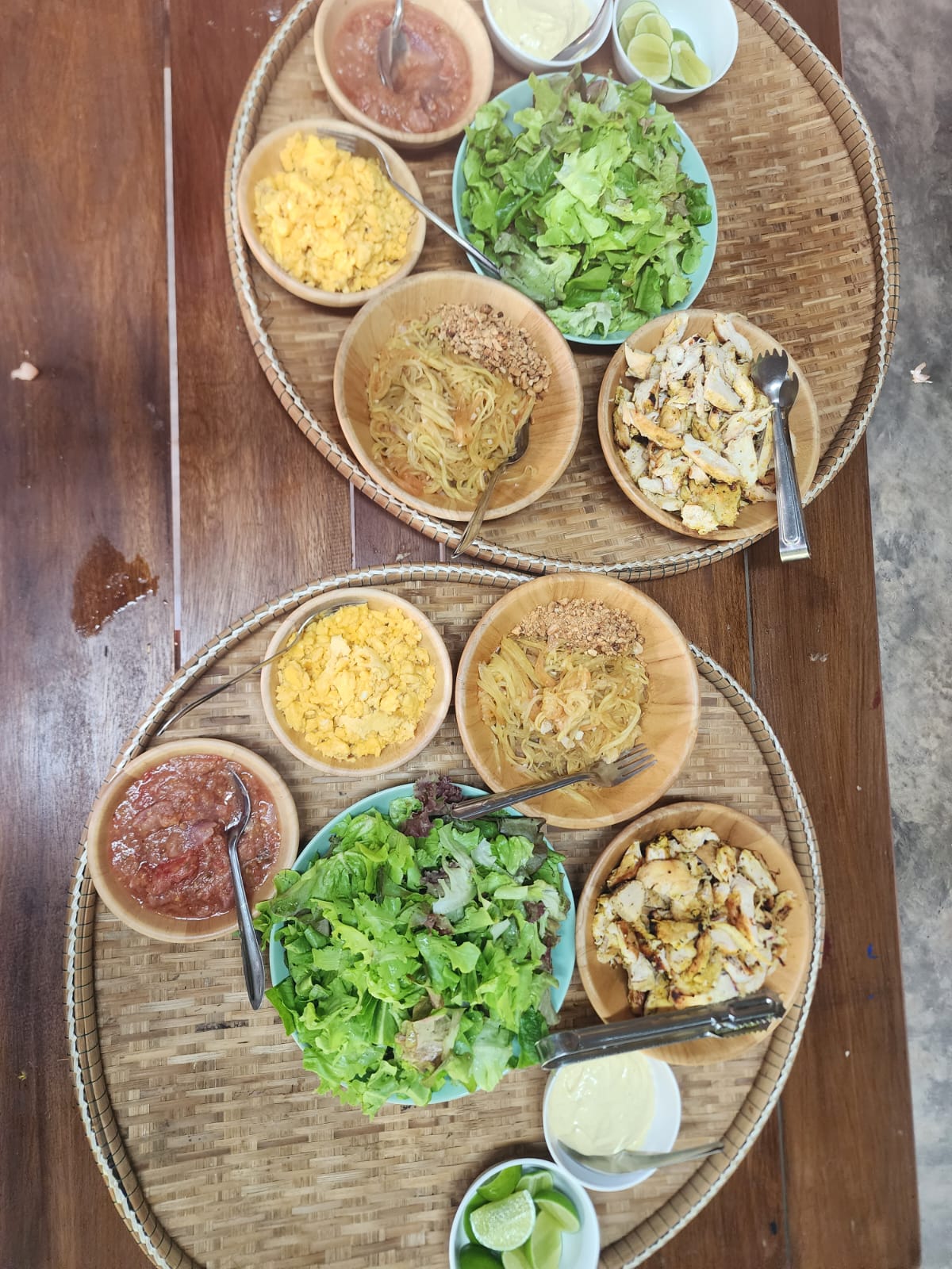 Woven trays hold salad, noodles, grilled chicken, and dipping sauces on a wooden table