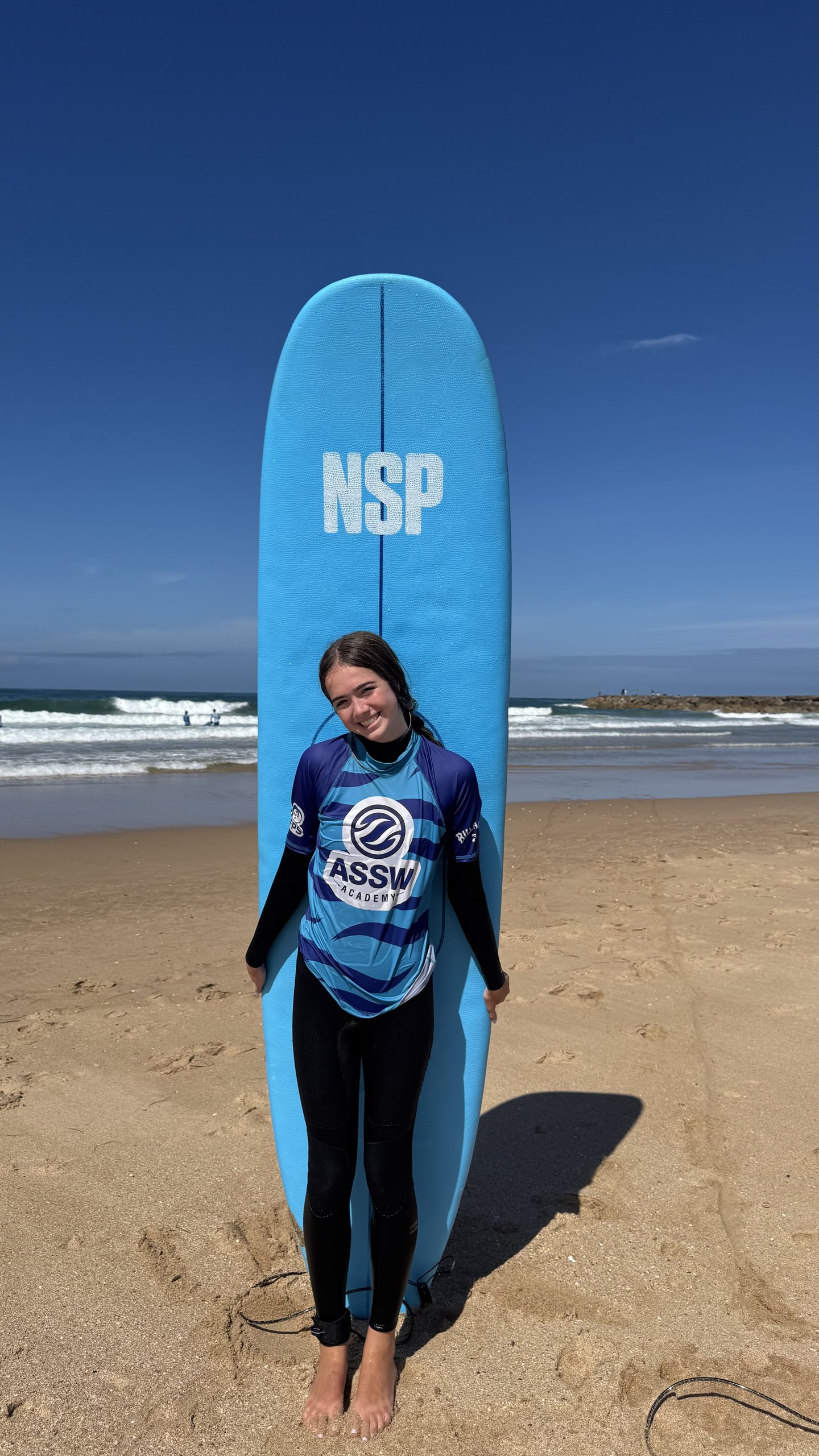 A teen in a surf rash guard stands beside a tall blue surfboard on a sandy beach