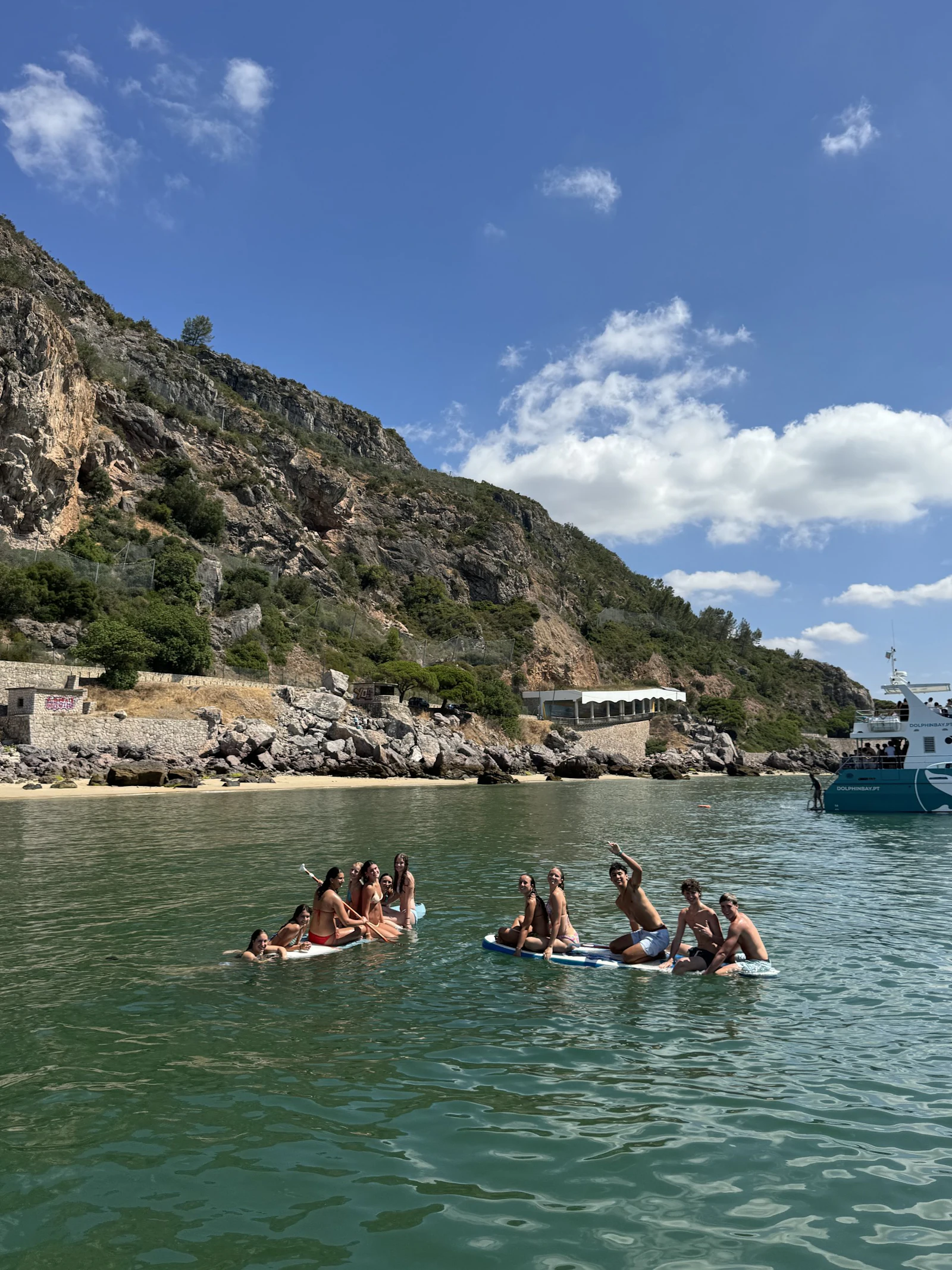 Teen travelers on paddleboards wave from a clear green cove below rocky cliffs