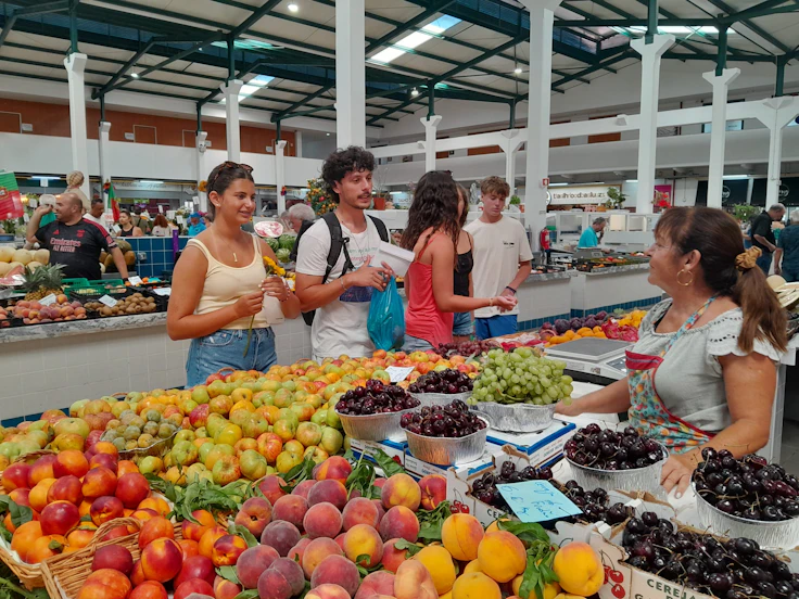 Teen travelers buy fresh fruit at a market stall piled with peaches, grapes and cherries