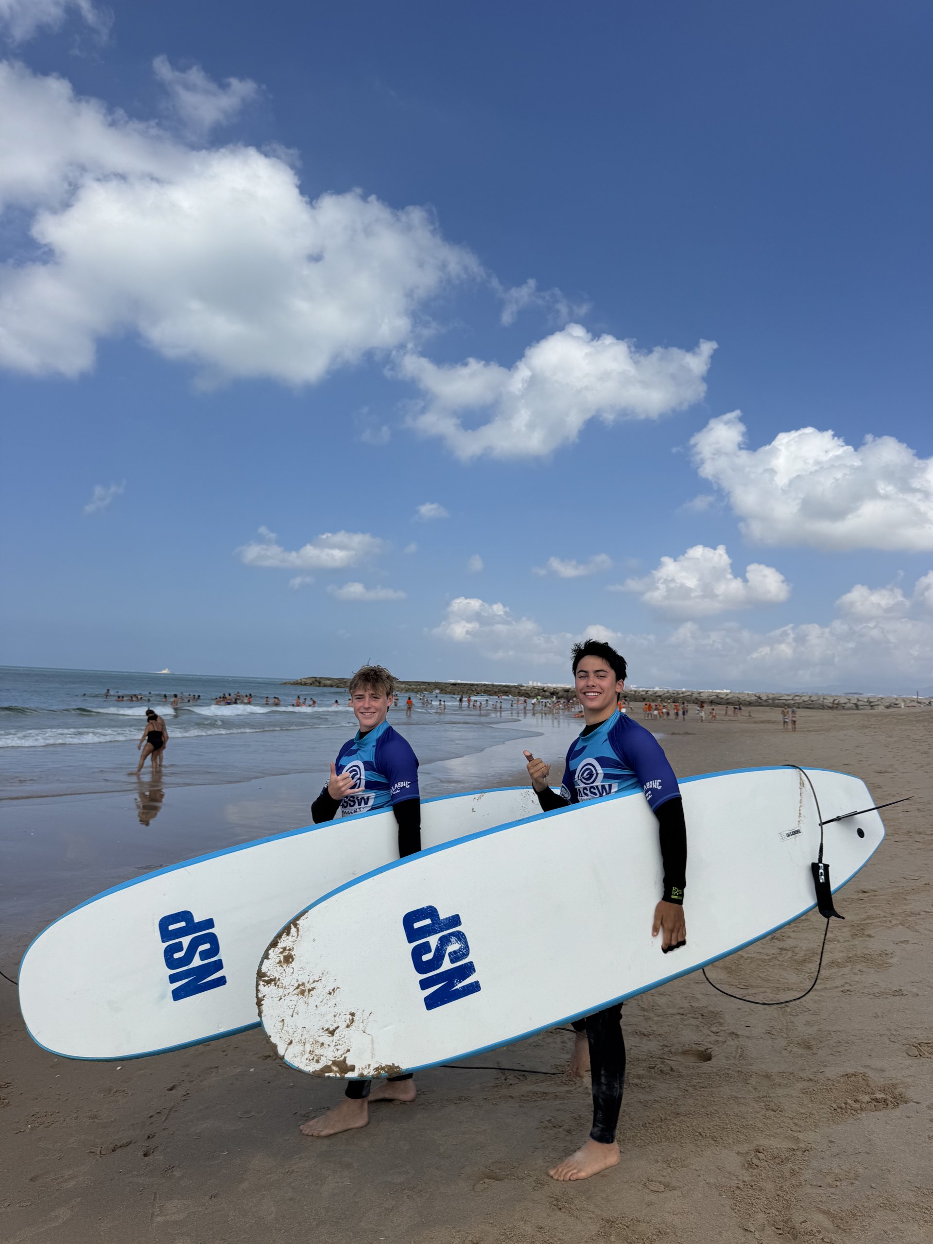 Two teens in wetsuits carry white surfboards across a wide sandy beach