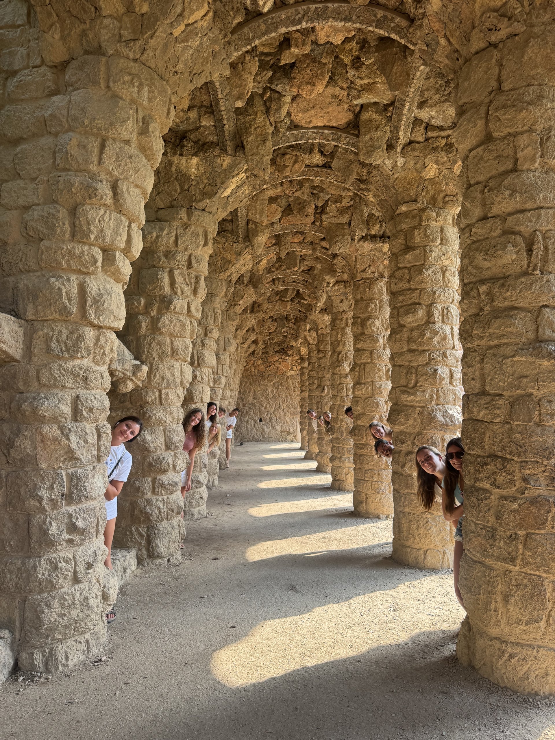 Teen travelers peek out from behind the stone columns of a covered walkway at Park Güell in Barcelona