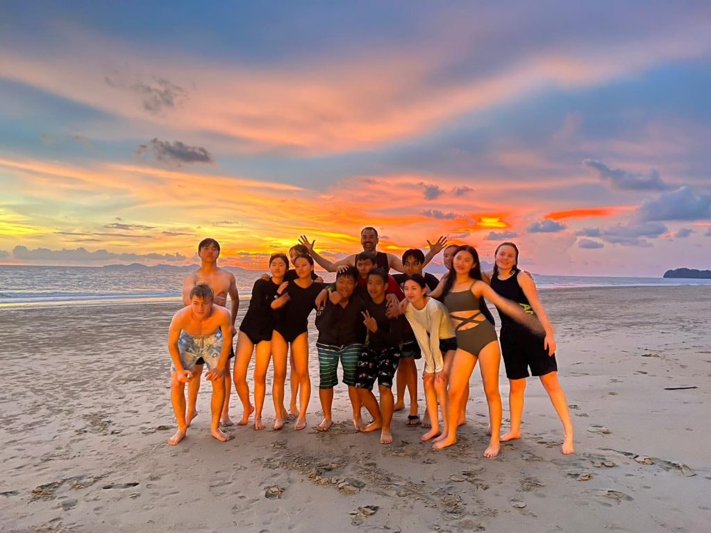 A group of students poses on a beach in Thailand while the sun is setting down.