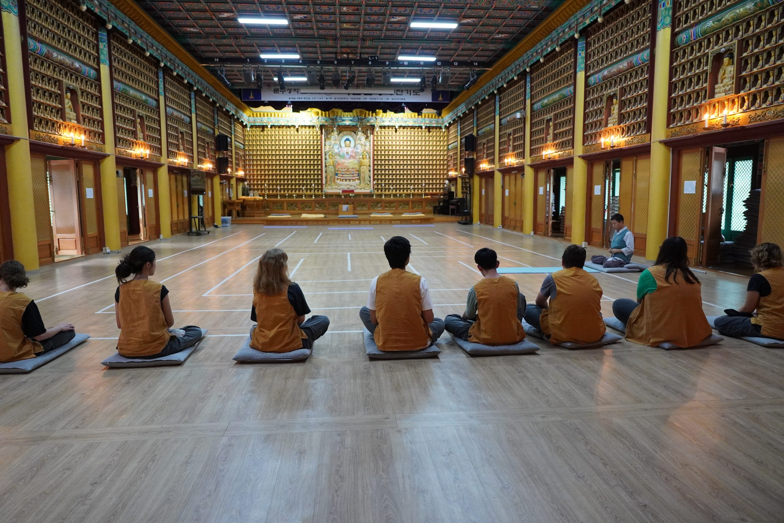 Students sit cross-legged in rows on the floor of a temple hall during a Buddhist practice session
