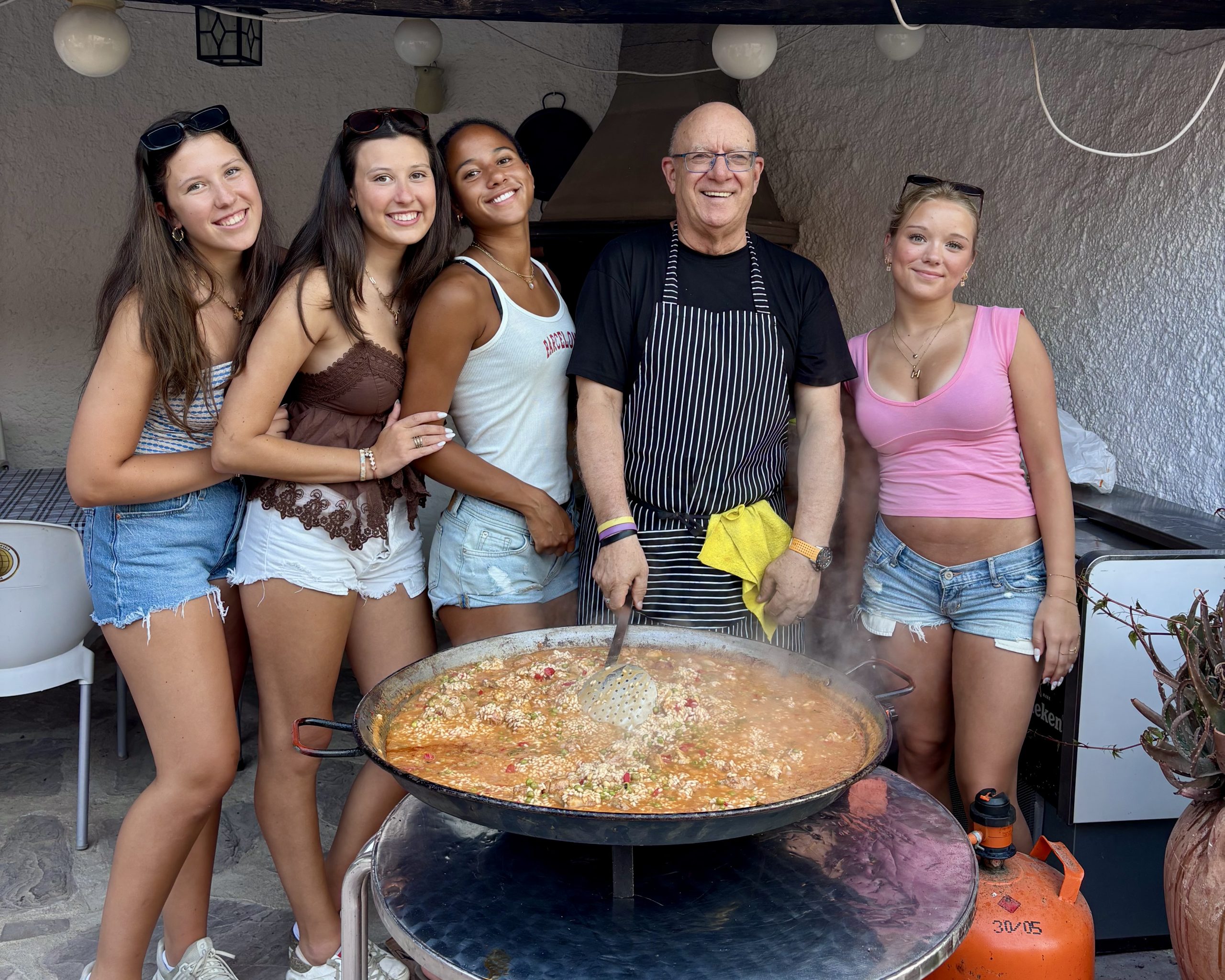 Students gather with a chef around a large pan of paella cooking outdoors