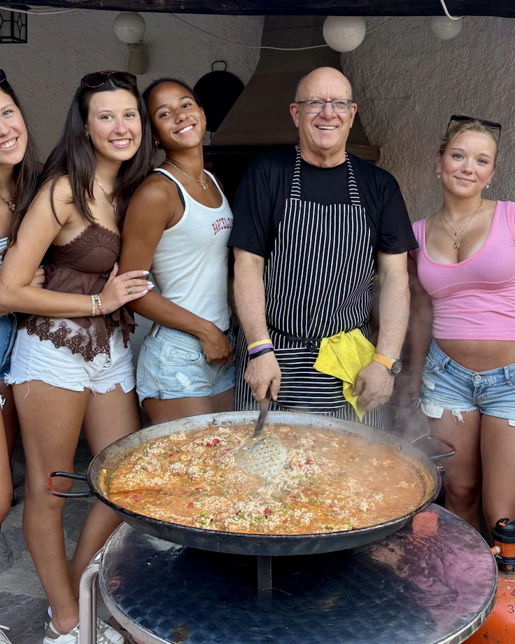 Four teen travelers pose with a chef in a striped apron beside a large pan of paella in Spain