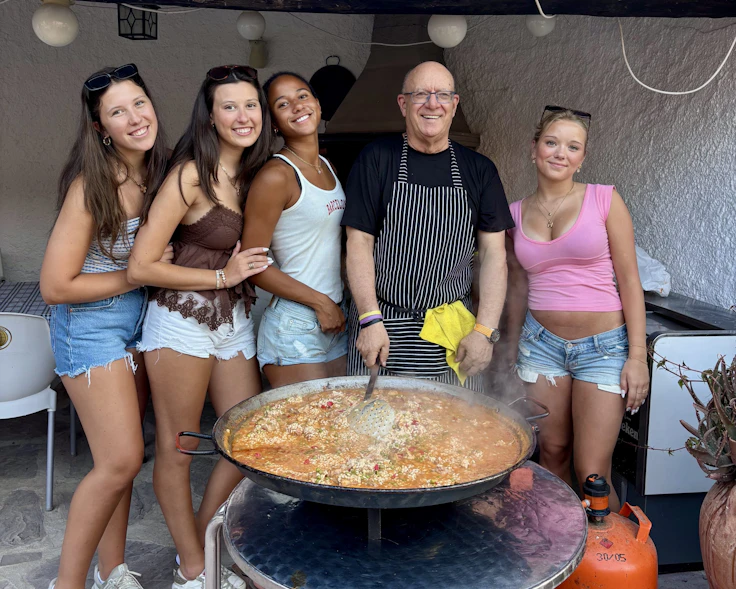 Four teen travelers pose with a chef in a striped apron beside a large pan of paella in Spain