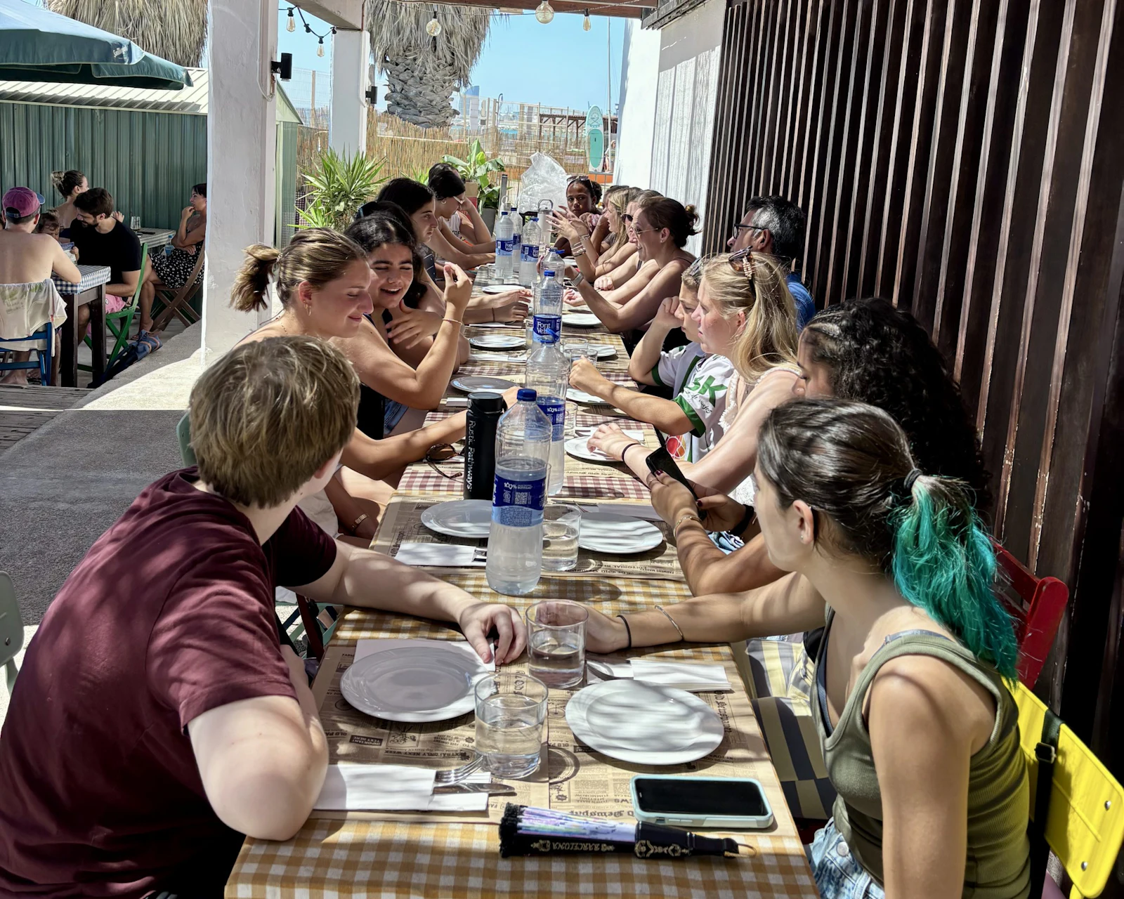Teen travelers share a meal at a long outdoor table under a shaded terrace in Spain
