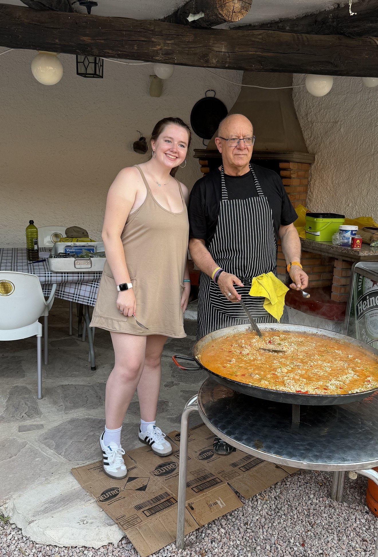 A teen traveler stands beside a chef in a striped apron stirring a large pan of paella in Spain