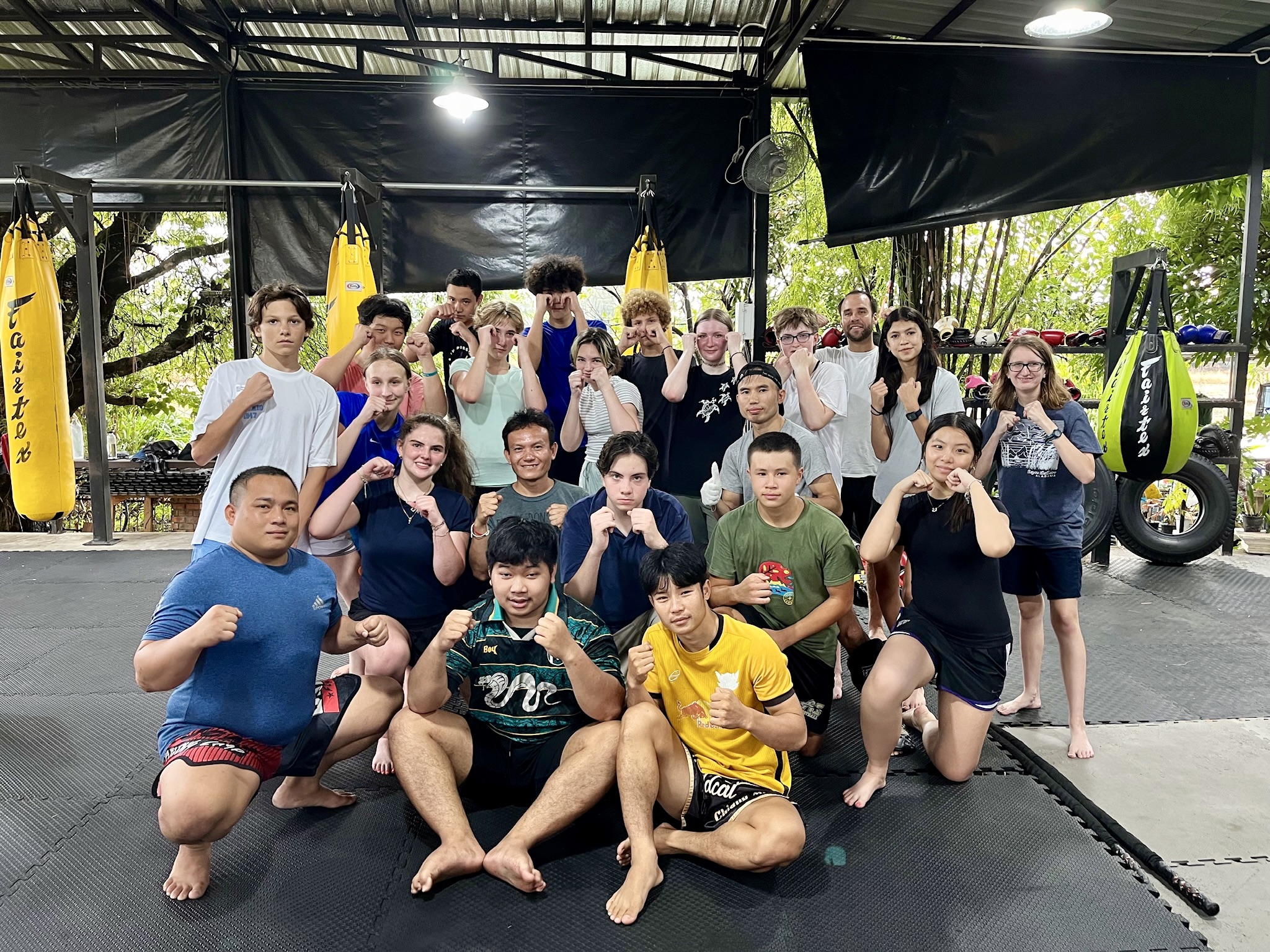 Students pose with raised fists at a Muay Thai gym under a covered training area