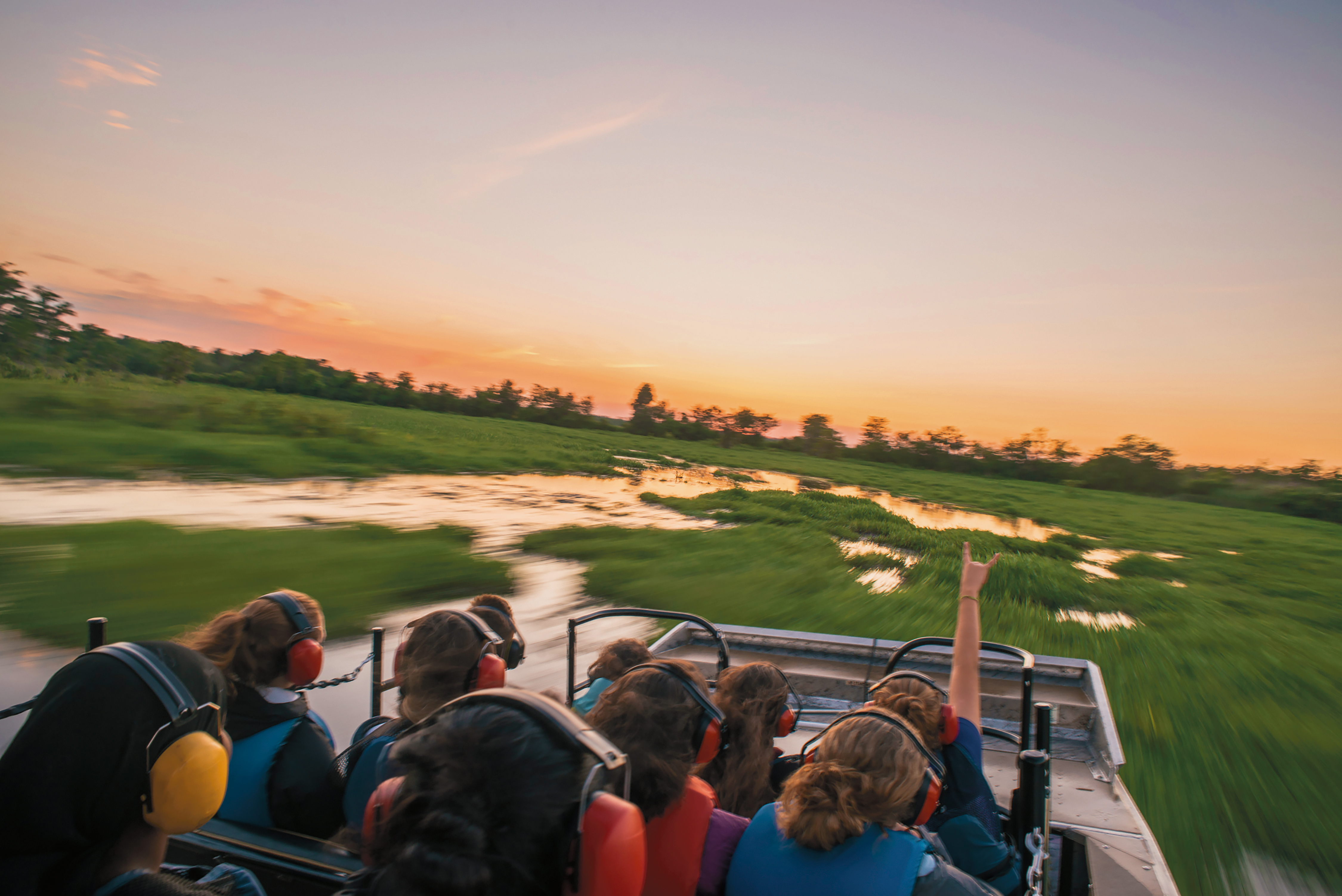 Students wear ear protection on an airboat speeding through the bayou at sunset