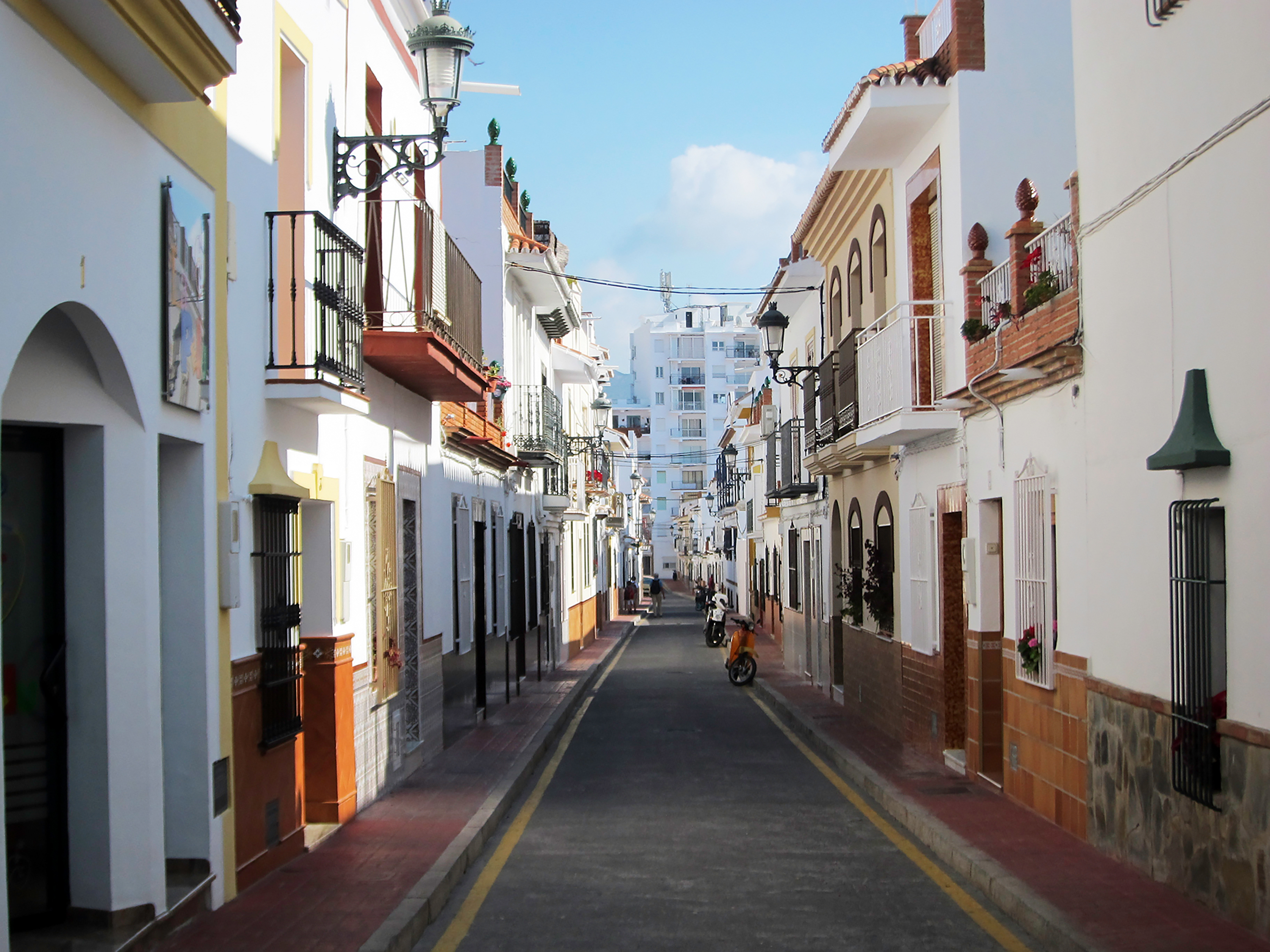 Students walk a narrow street lined with whitewashed buildings and wrought-iron balconies in a Spanish town