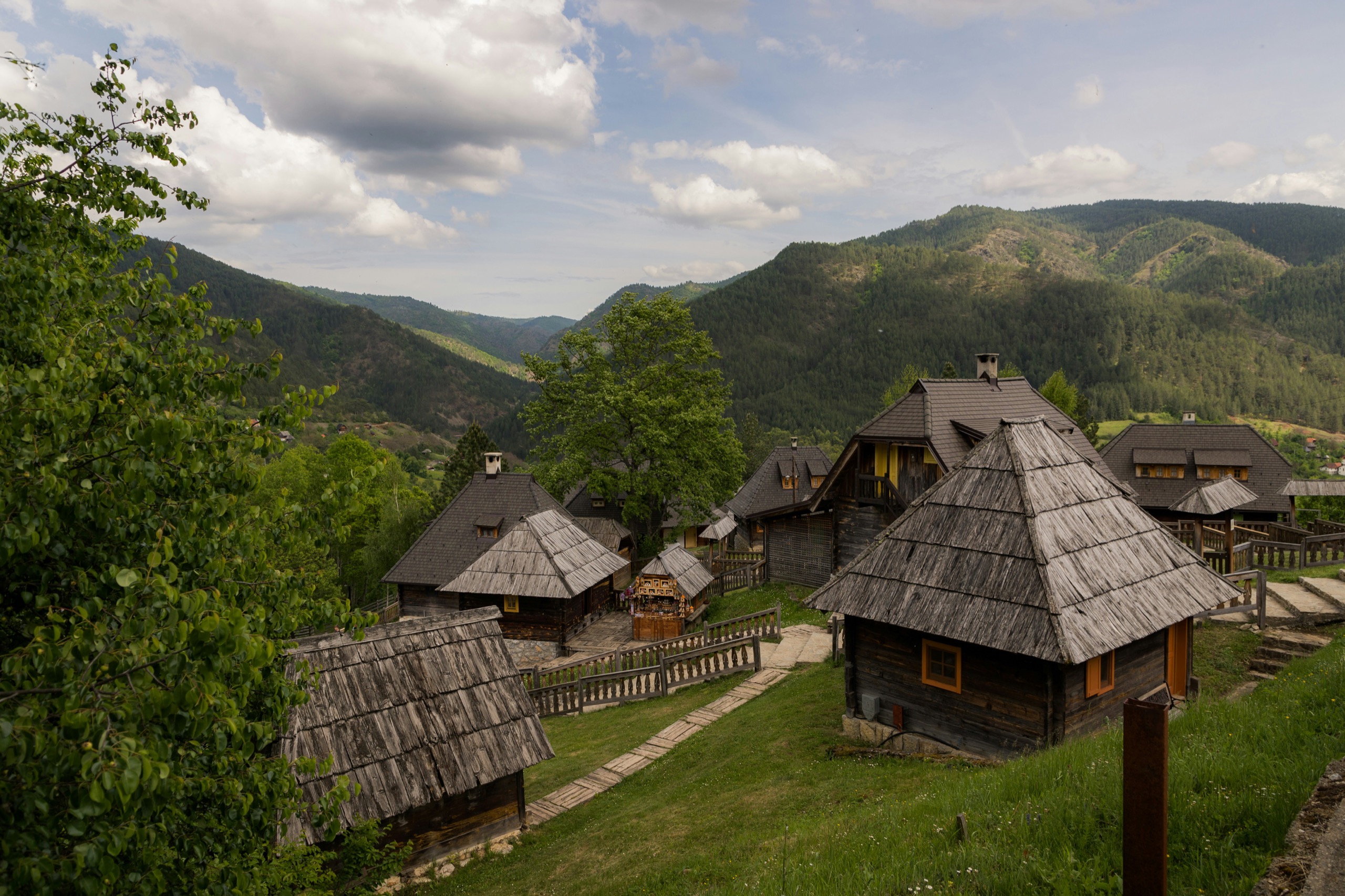 Traditional wooden houses with shingled roofs cluster along a fenced path in a forested mountain village.