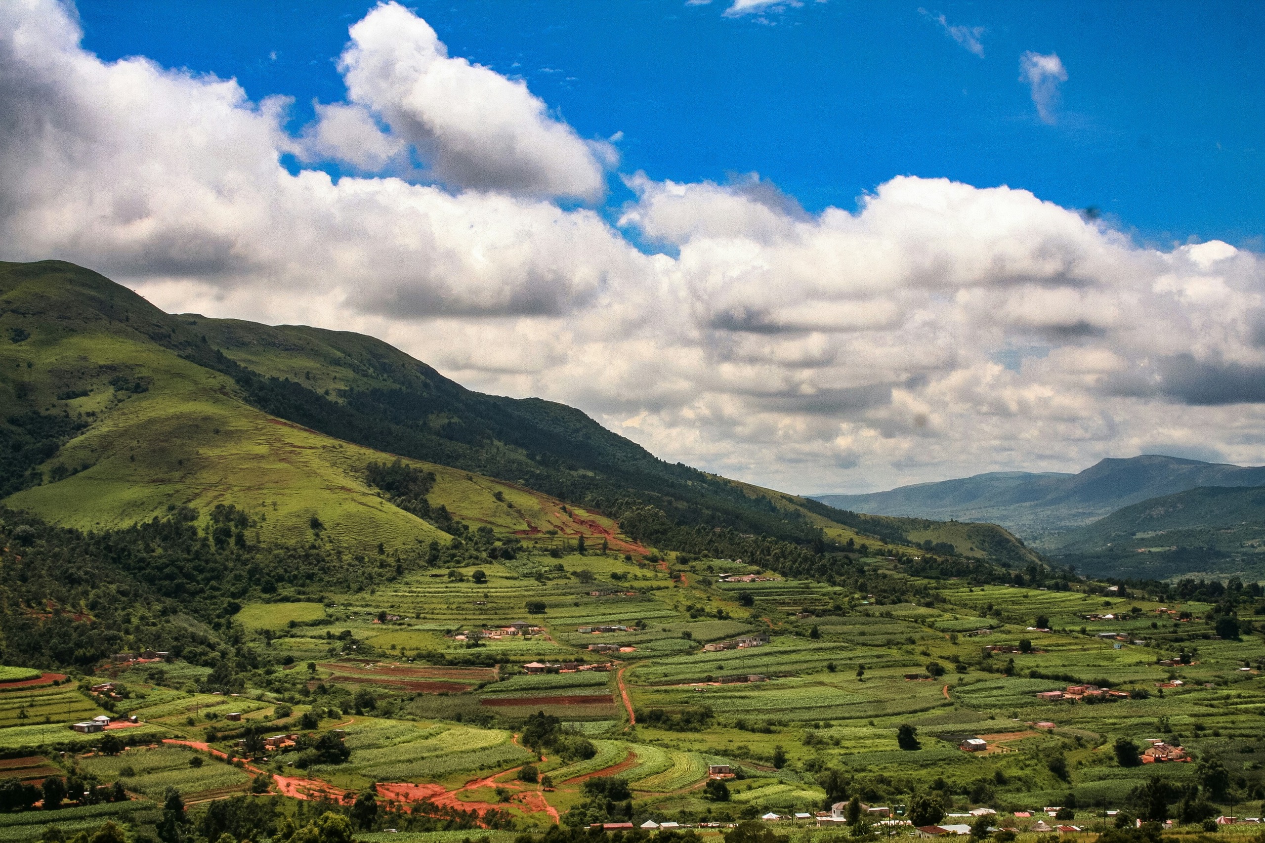 Green hills in Eswatini fall to a valley of terraced fields and scattered homesteads, with heaped white clouds over the ridges.