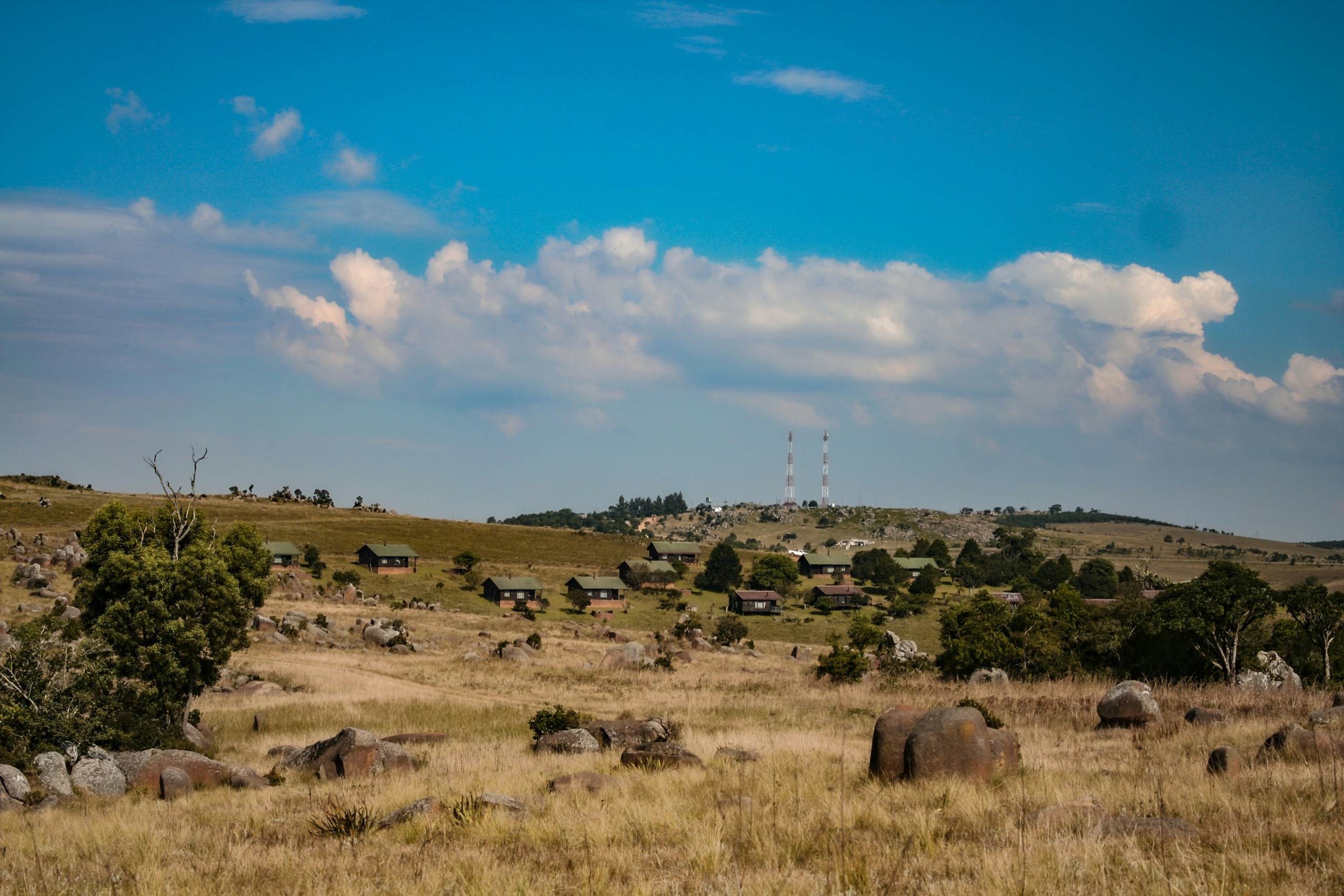 Granite boulders scattered across dry highland grassland, with a few thatched buildings and two radio masts on the ridge under a blue sky.