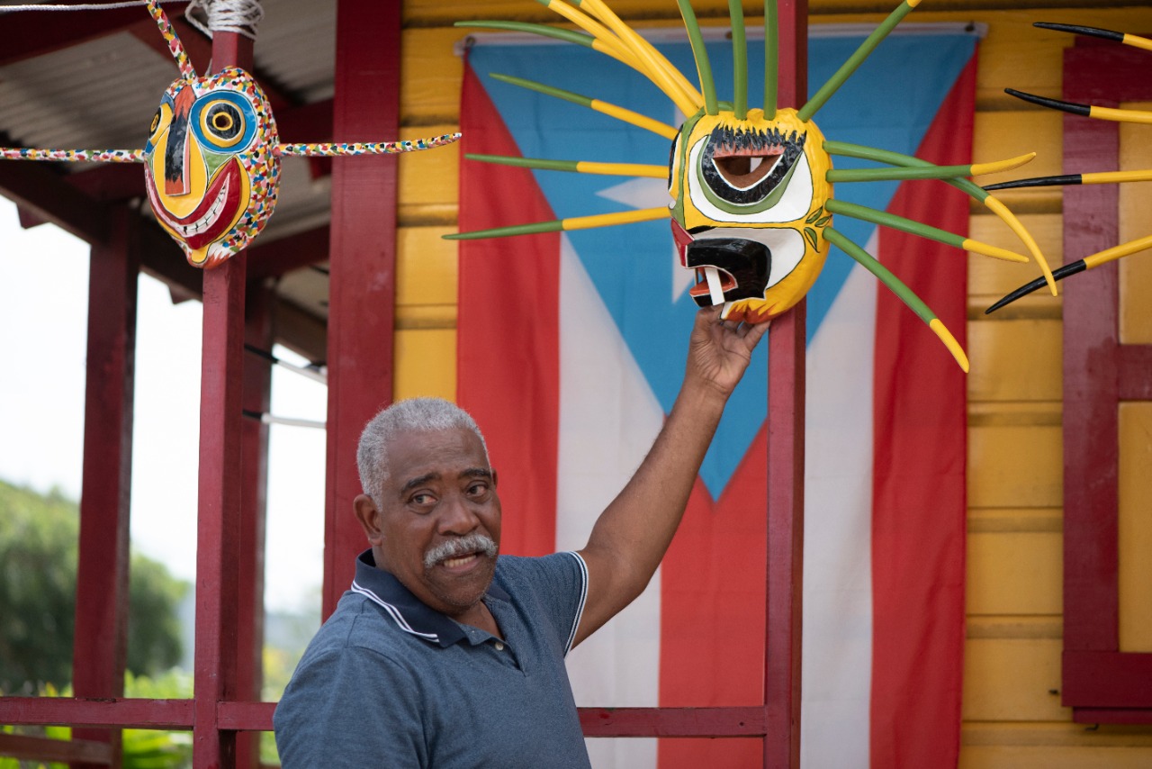 Man holding a traditional Puerto Rican mask with flag in the background