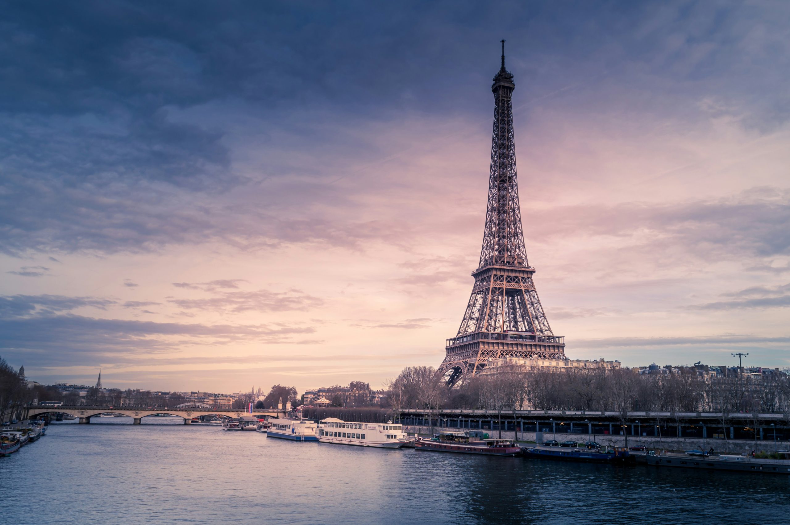 The Eiffel Tower rises against a pink and blue sunset sky, seen across the Seine with tour boats moored along the riverbank.