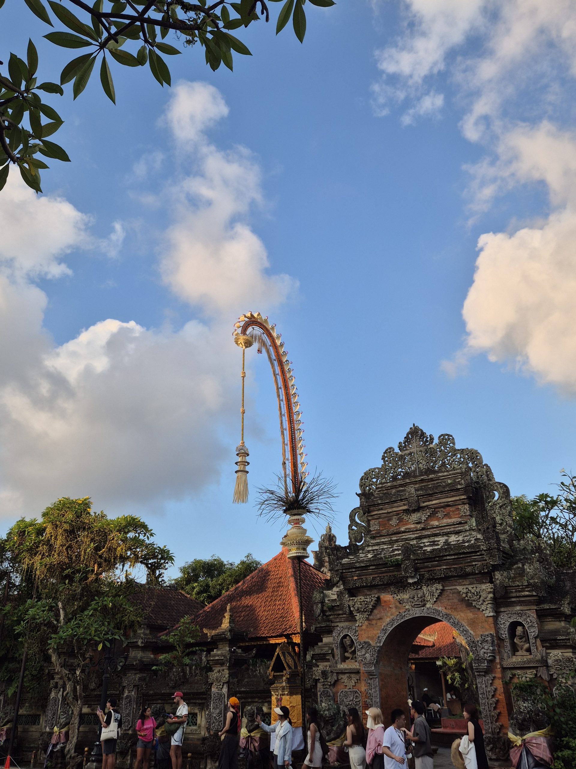 A tall, curved bamboo penjor decoration arches above an ornately carved stone temple gateway, framed by tree branches against a partly cloudy sky.