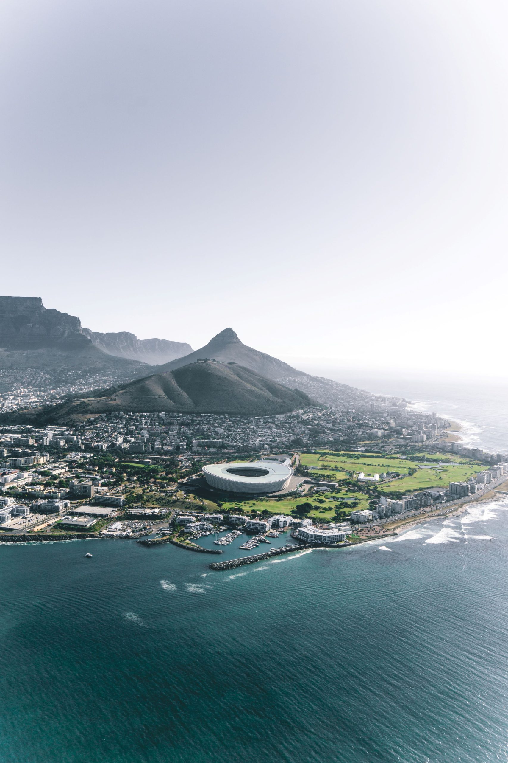 An aerial view along a city built between mountains and the sea, with a white stadium and a harbour on the shoreline, and a peaked mountain rising behind the streets.