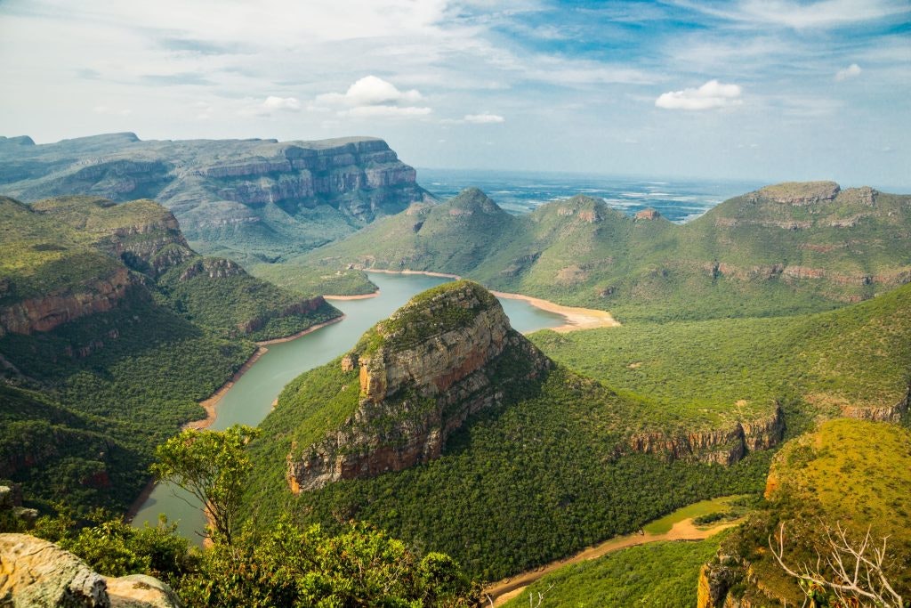 This image shows a stunning landscape of lush green mountains surrounding a winding river. The view features dramatic cliffs and valleys, creating a peaceful yet striking natural scene.The location is the Blyde River Canyon in South Africa, one of the largest canyons in the world. It's known for its rich biodiversity and beautiful views like this one.