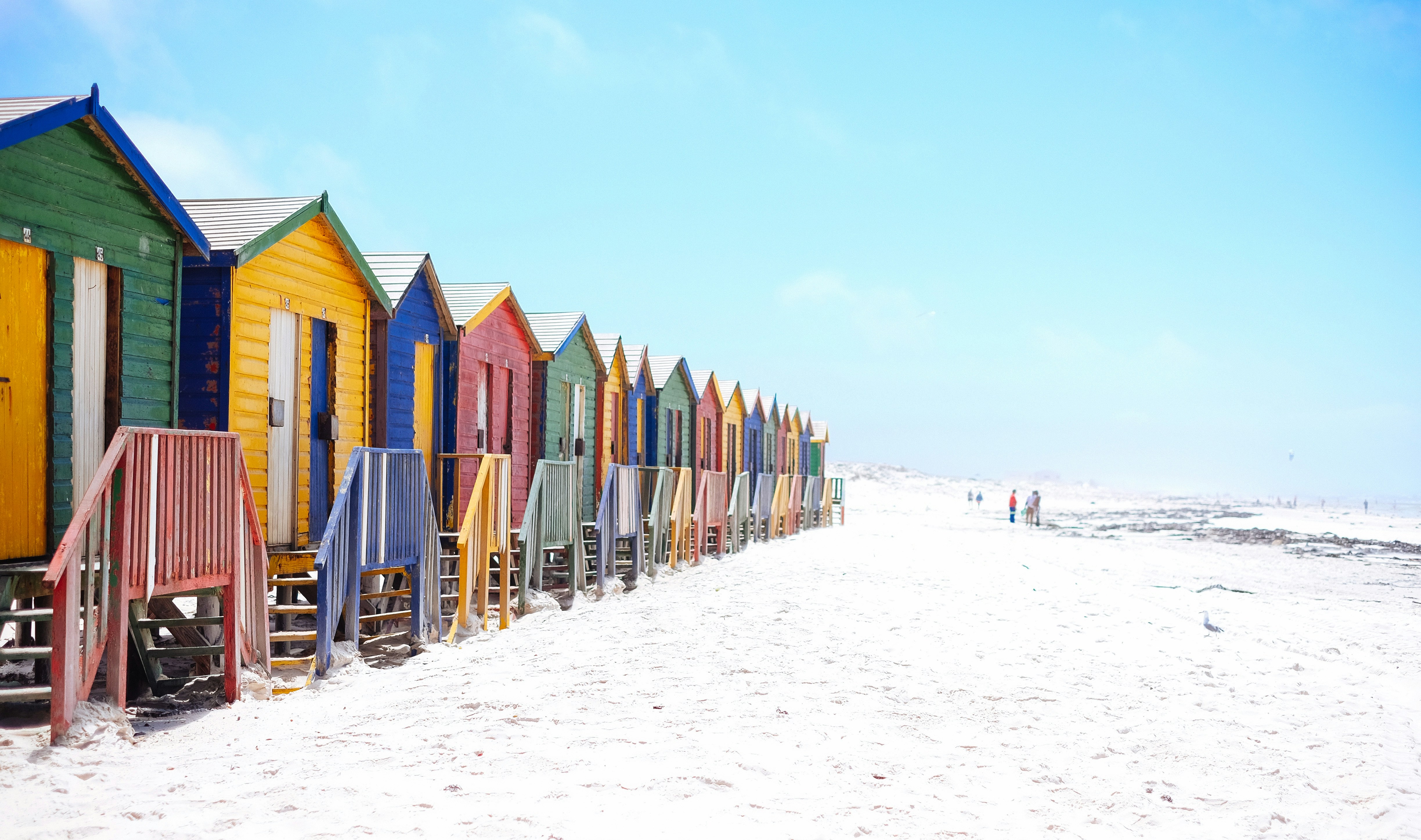 A row of brightly painted wooden beach huts in green, yellow, blue, and red runs along white sand at St James Beach, with a few people walking at the water's edge under a clear sky.