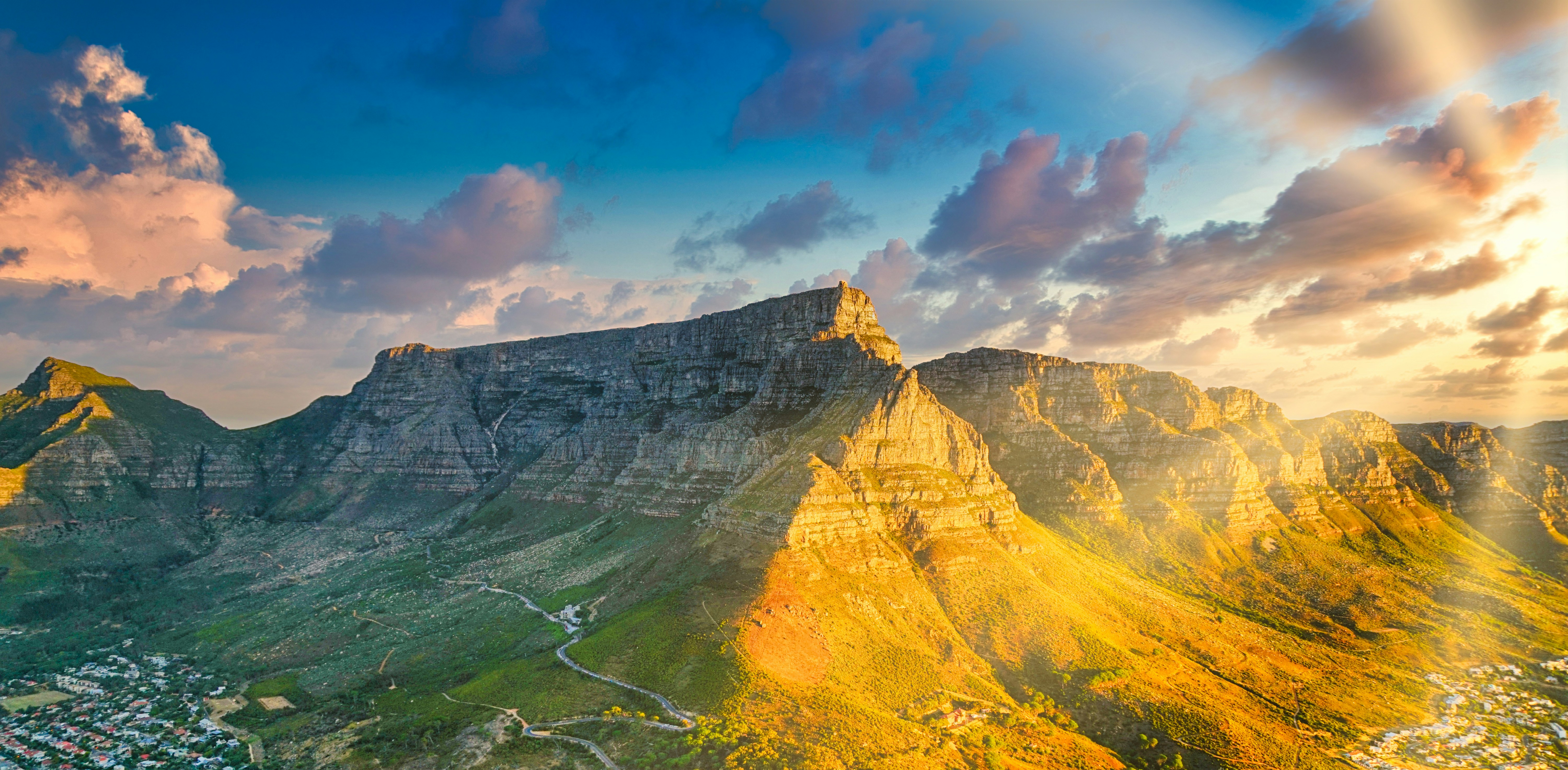 Table Mountain's flat sandstone summit catches low evening sun, with a road winding across its green lower slopes and houses at its foot.