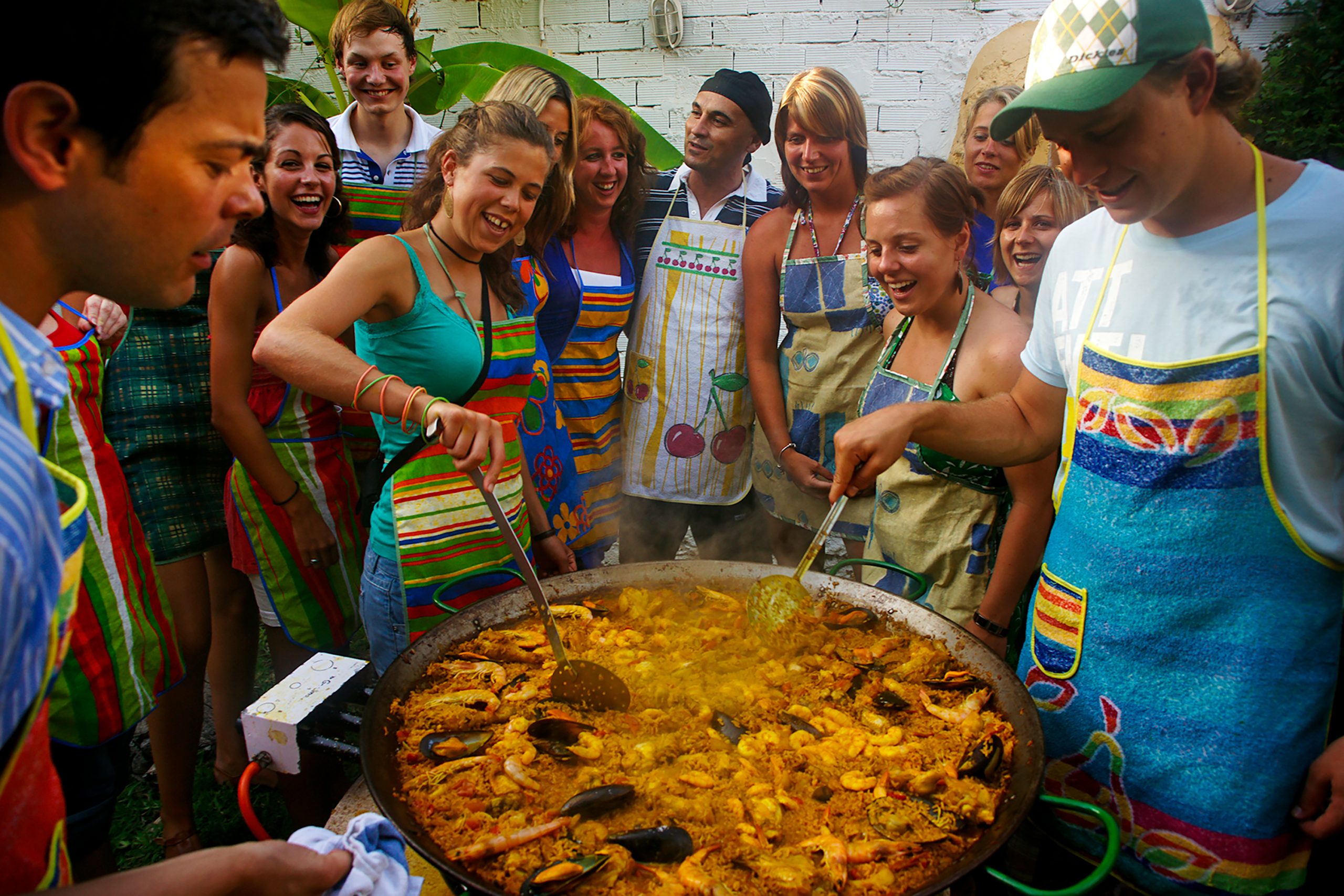 Students in aprons gather around a large pan of paella cooking outdoors in Spain