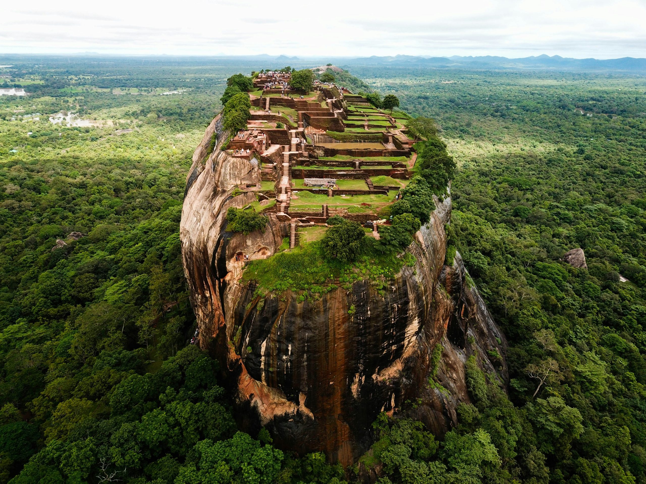 An aerial view of Sigiriya rock fortress, with the terraced ruins of the summit palace above the surrounding forest.