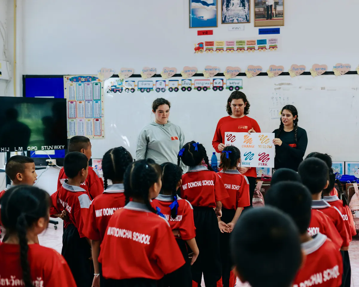 Three students standing at the front of a Thai classroom holding a colour chart while a class of children in red uniforms watches