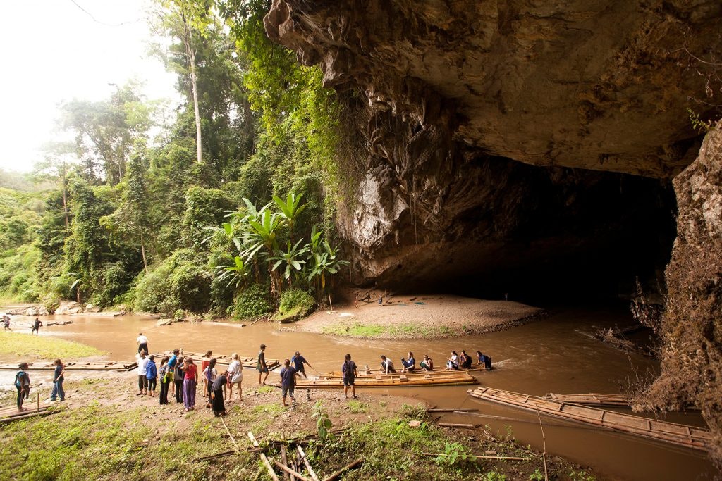 Group of teens entering a large limestone cave near a river in Thailand, surrounded by lush greenery.