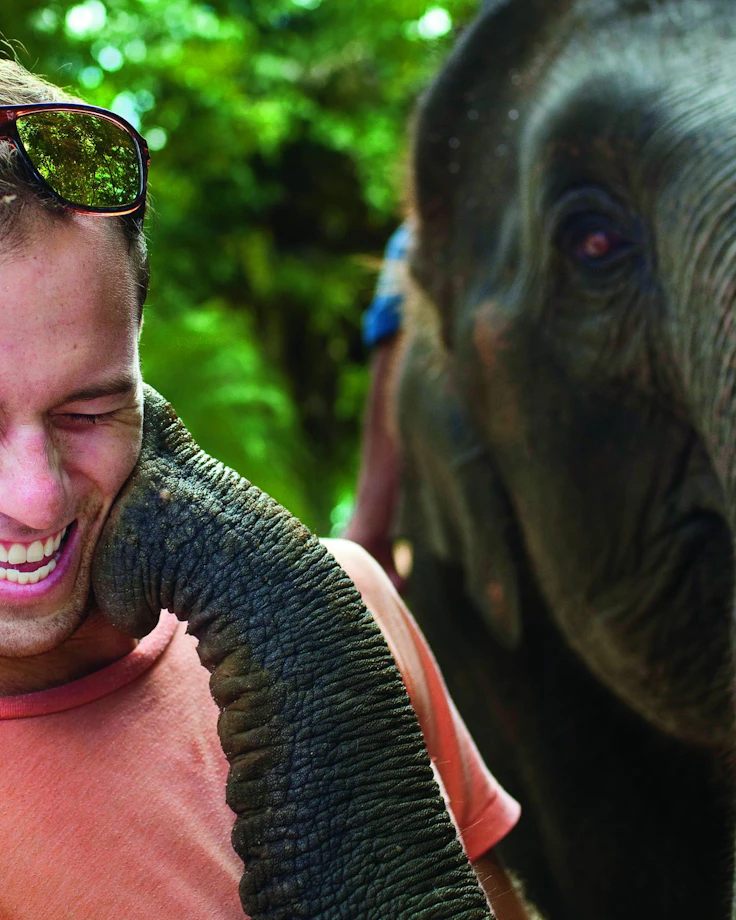 A laughing teen traveler in sunglasses has an elephant resting its trunk on their shoulder at an elephant sanctuary.