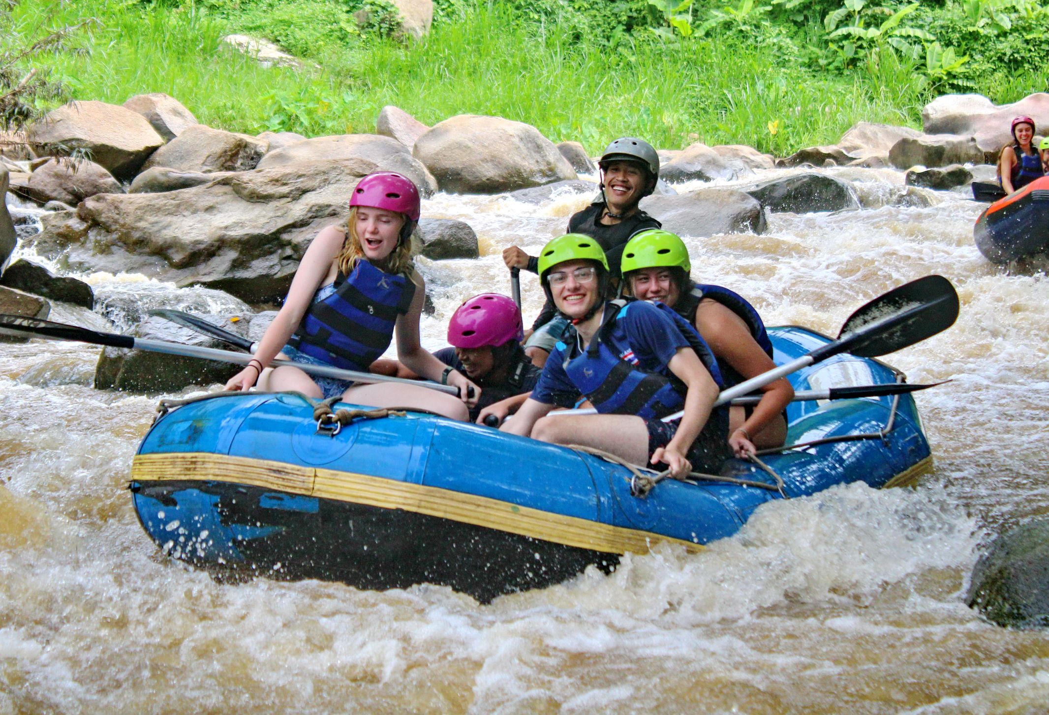 Students wearing helmets and life jackets paddle a raft through rapids